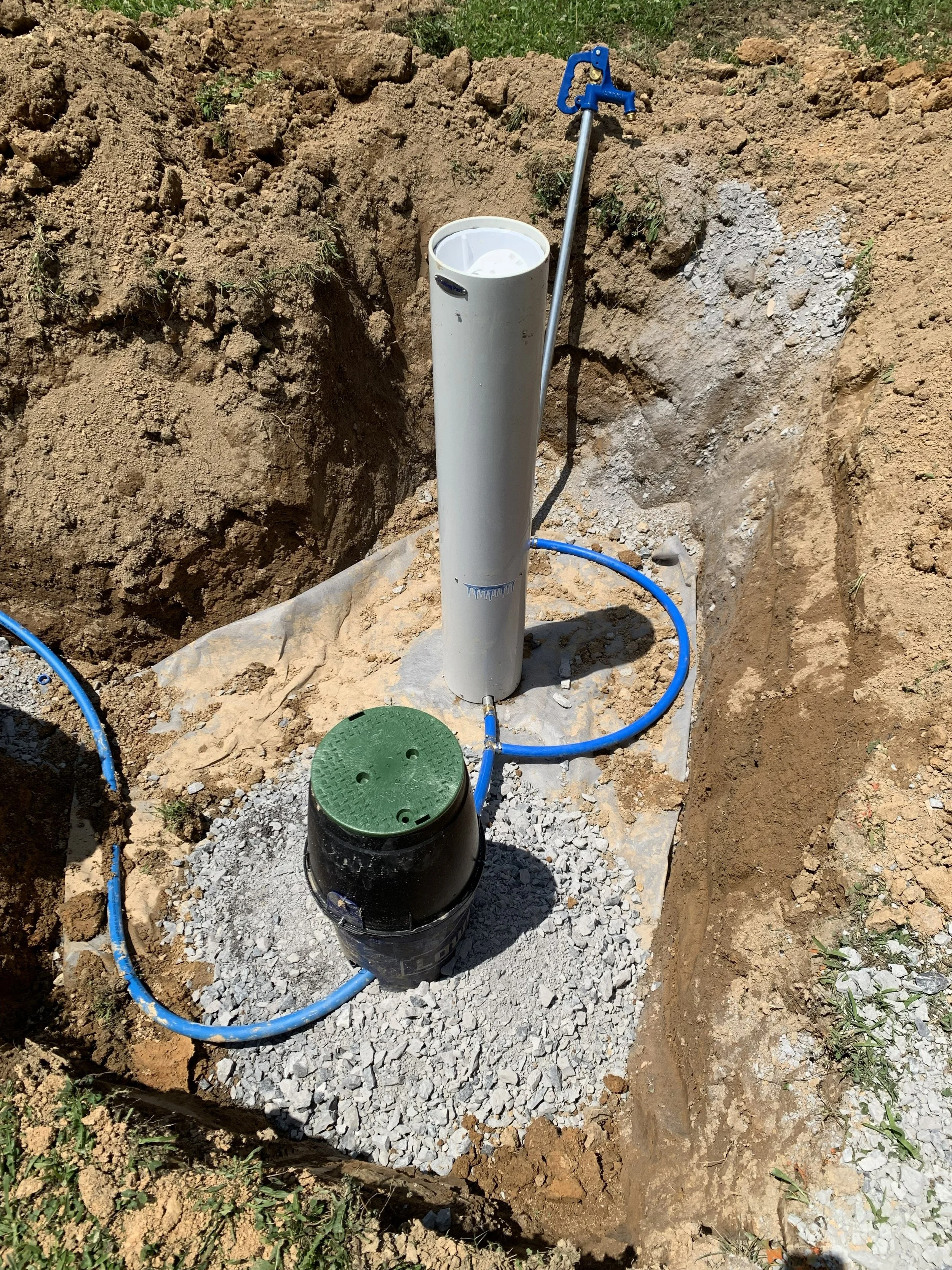 An underground utility access pipe with a white pipe and blue piping, set in a dug-out area with dirt walls and rocky gravel base, and a green covered container nearby.