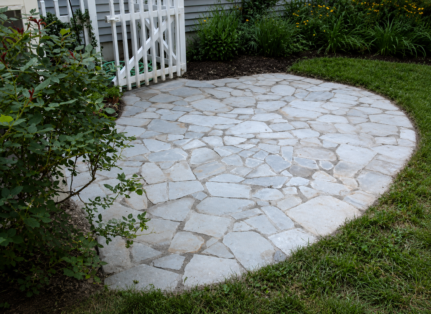 A backyard patio made of irregularly shaped light-colored stones, surrounded by green grass and plants, with a white wooden fence and gardening hose in the background.