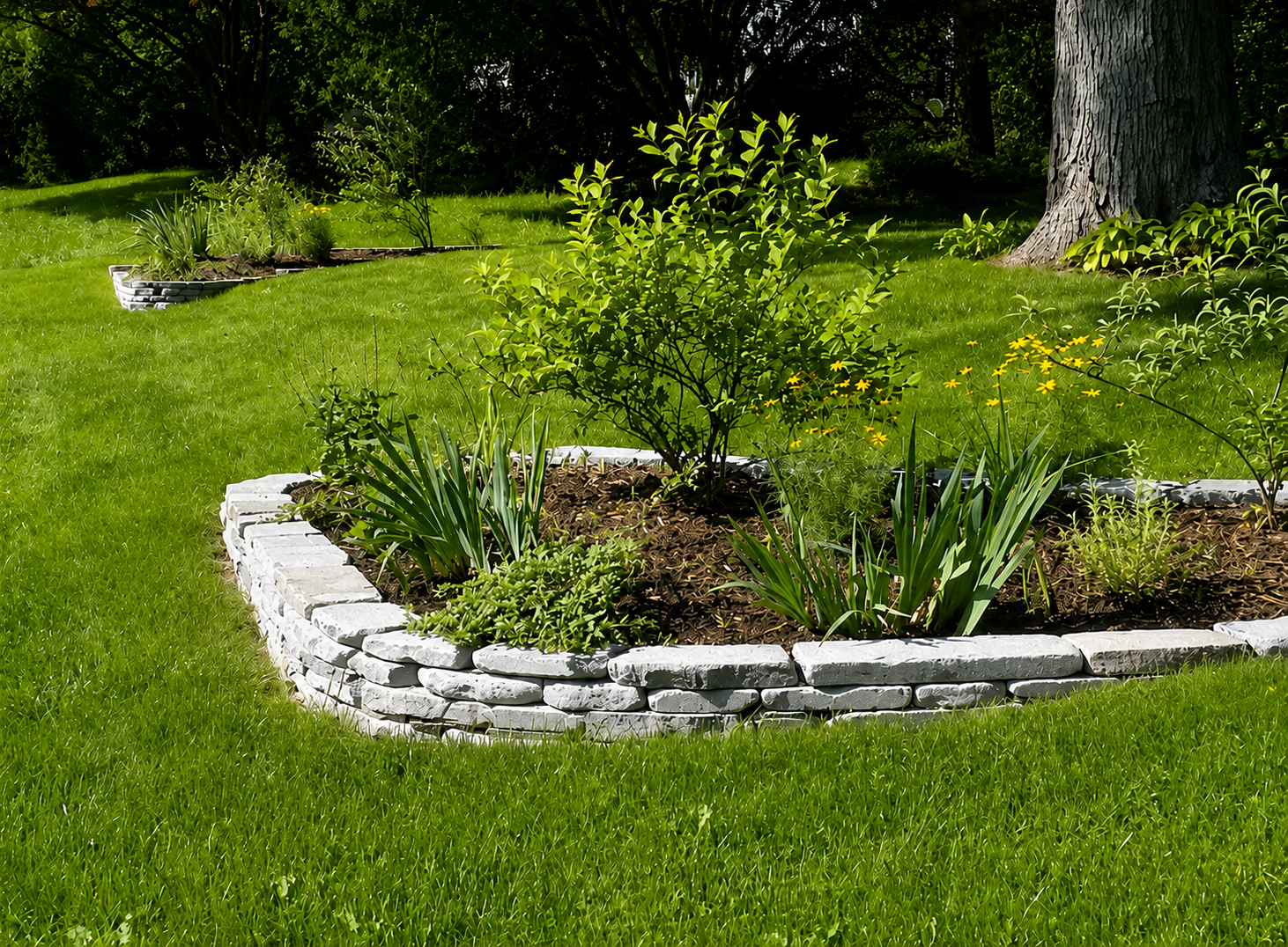 A landscaped garden bed with green plants and small yellow flowers, bordered by white bricks, on a lush green lawn with trees in the background.