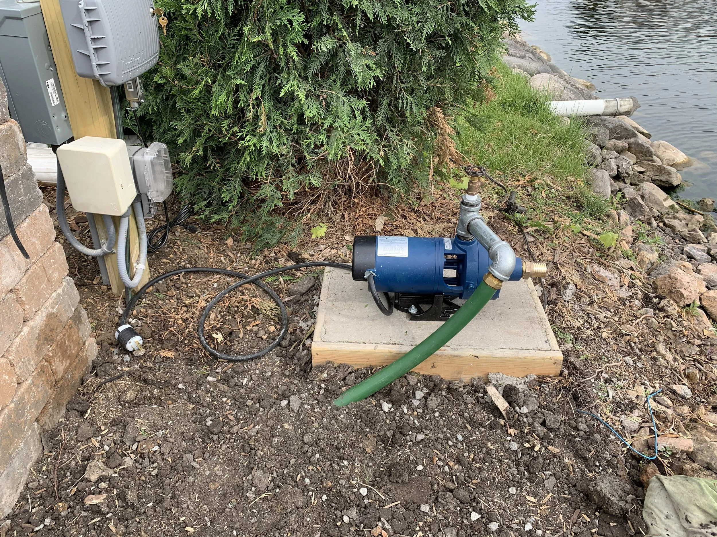 A small outdoor setup near a body of water with electronic equipment and a water pump connected to a green hose, surrounded by rocks and plants.