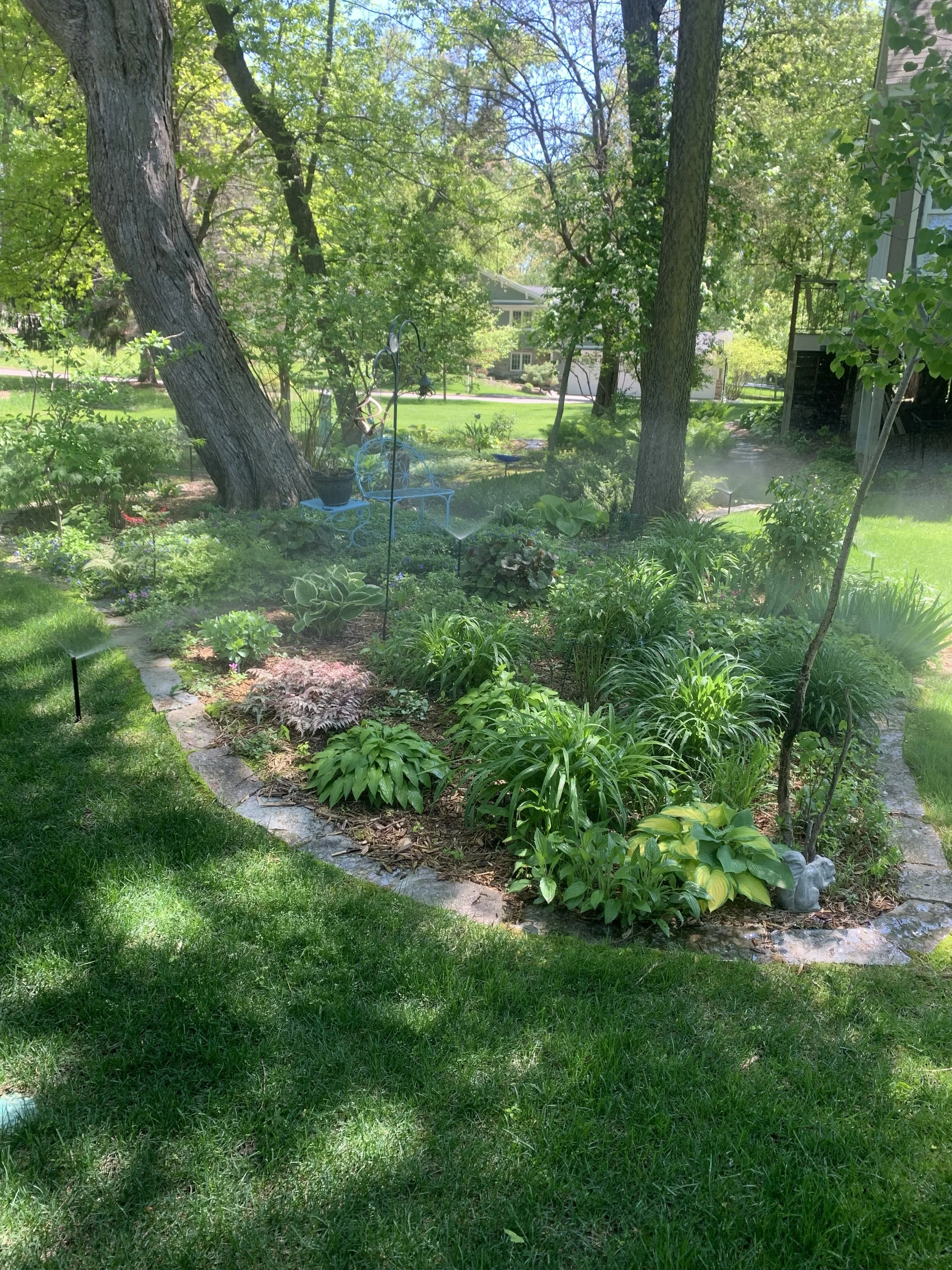 A lush backyard garden with green grass, various plants, trees, and a patio area with a white wrought iron bench and decorative garden accessories, basking in bright sunlight.
