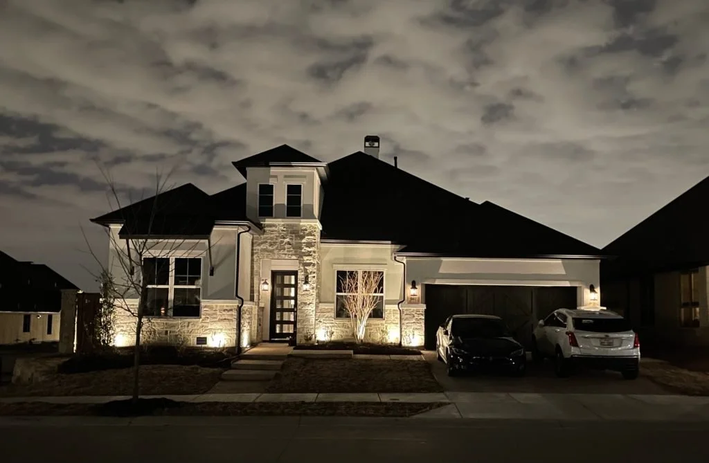 A modern two-story house at night with exterior lights illuminating the front, showing a stone and stucco facade, a driveway with two cars, and a leafless tree in the front yard.