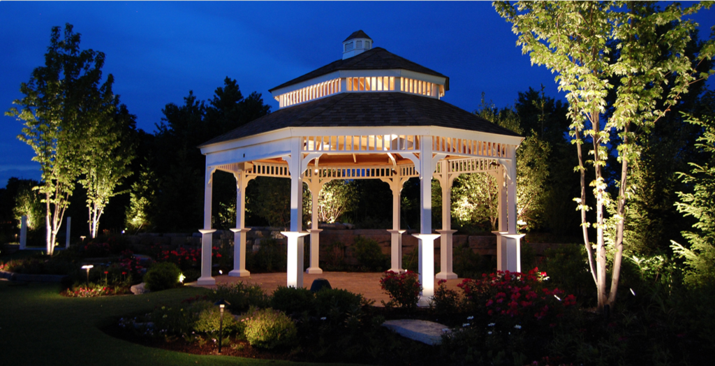 A white gazebo with decorative trim illuminated at night, surrounded by greenery and flowering plants.