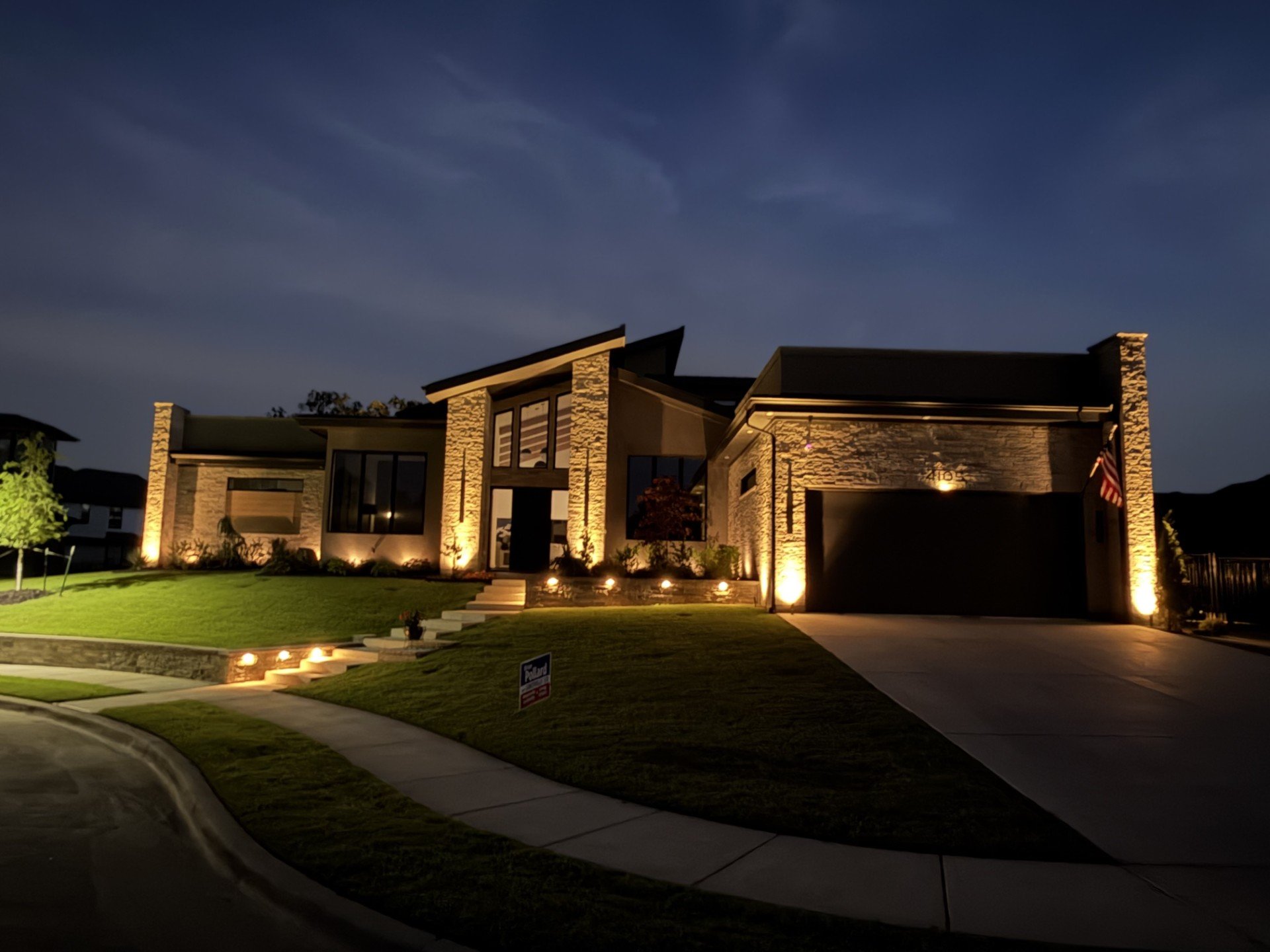 Modern house with stone facade and large windows illuminated at night, front yard with lawn and pathway lights, American flag near the garage.