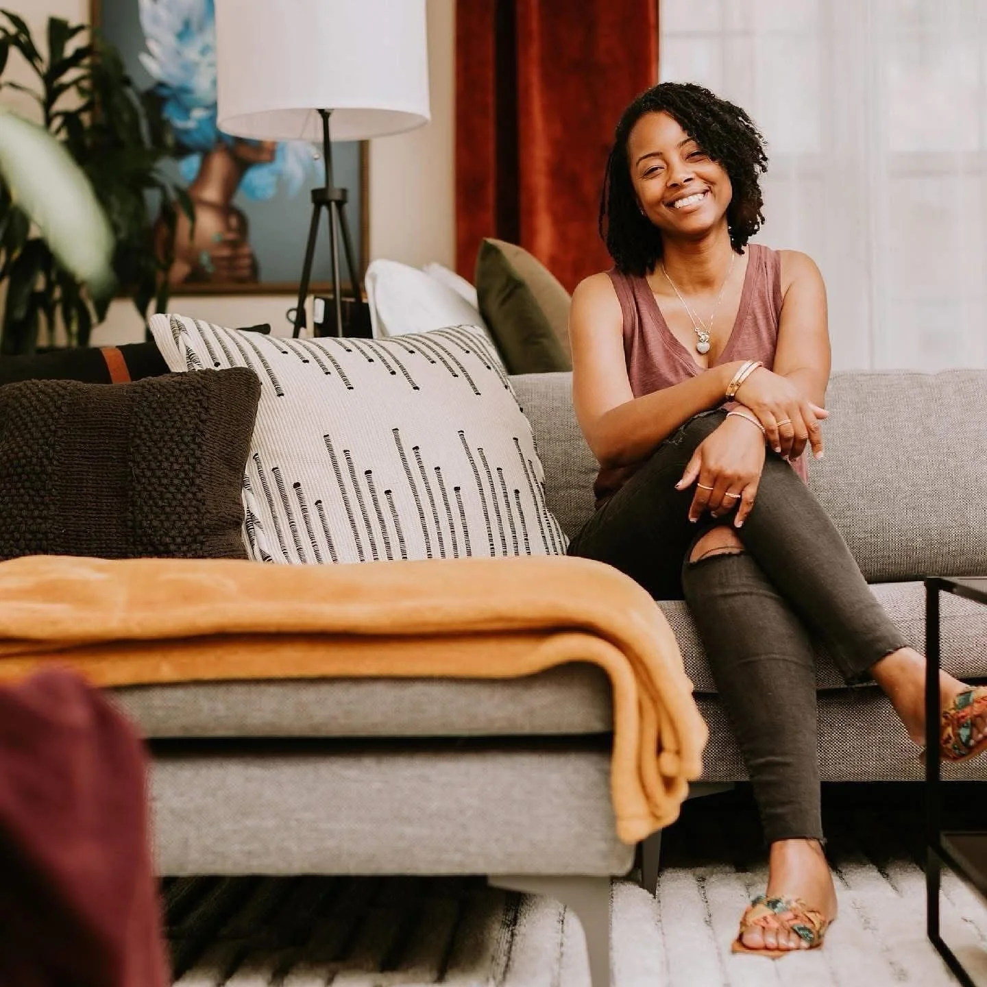 A woman sitting on a gray sofa, smiling, with various patterned pillows and a blanket in a cozy living room.