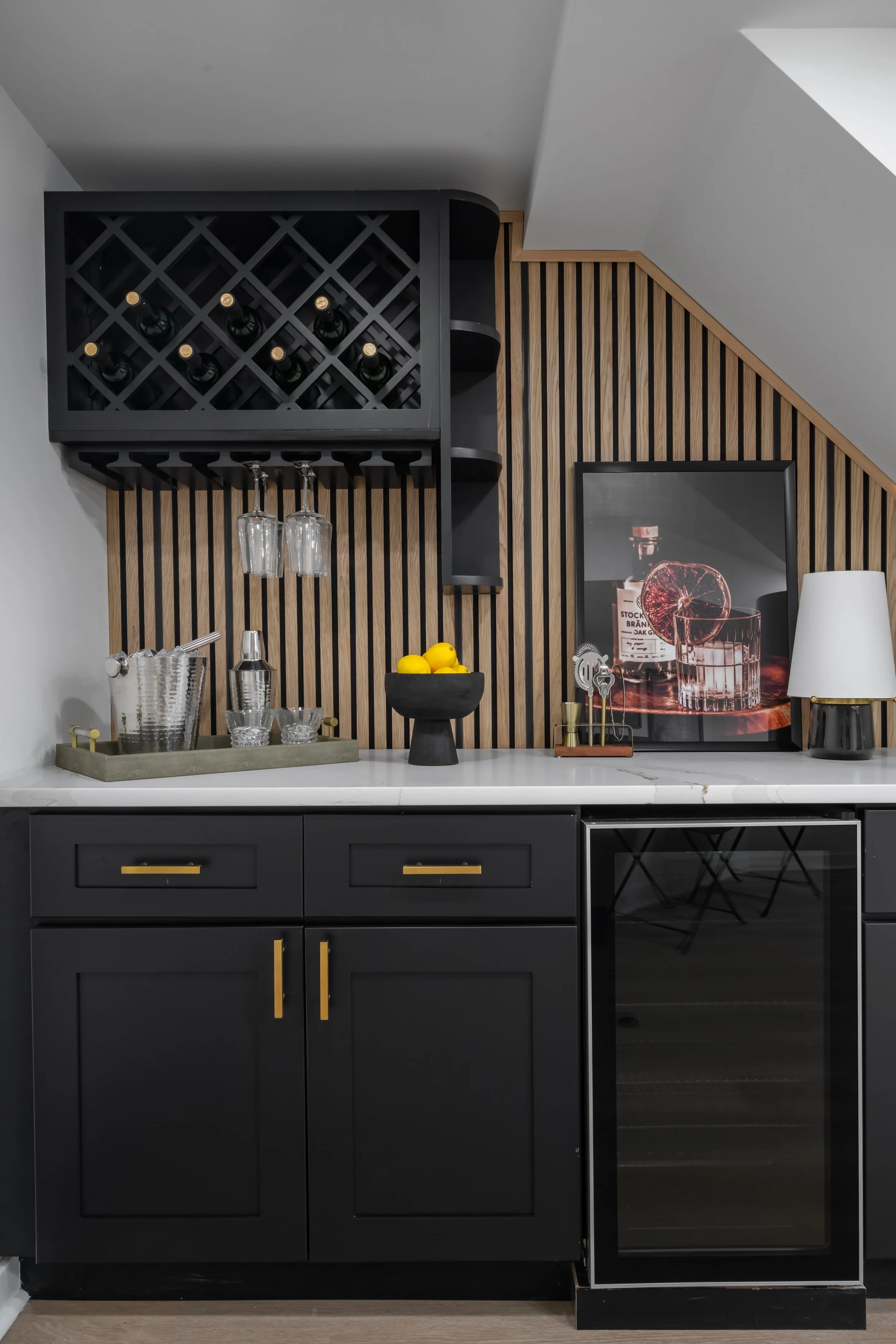 Black kitchen cabinet with gold handles, a white marble countertop, a black wine rack with bottles, hanging wine glasses, a framed photo of a cocktail with lemon slices, and decorative items including lemons and a lamp.