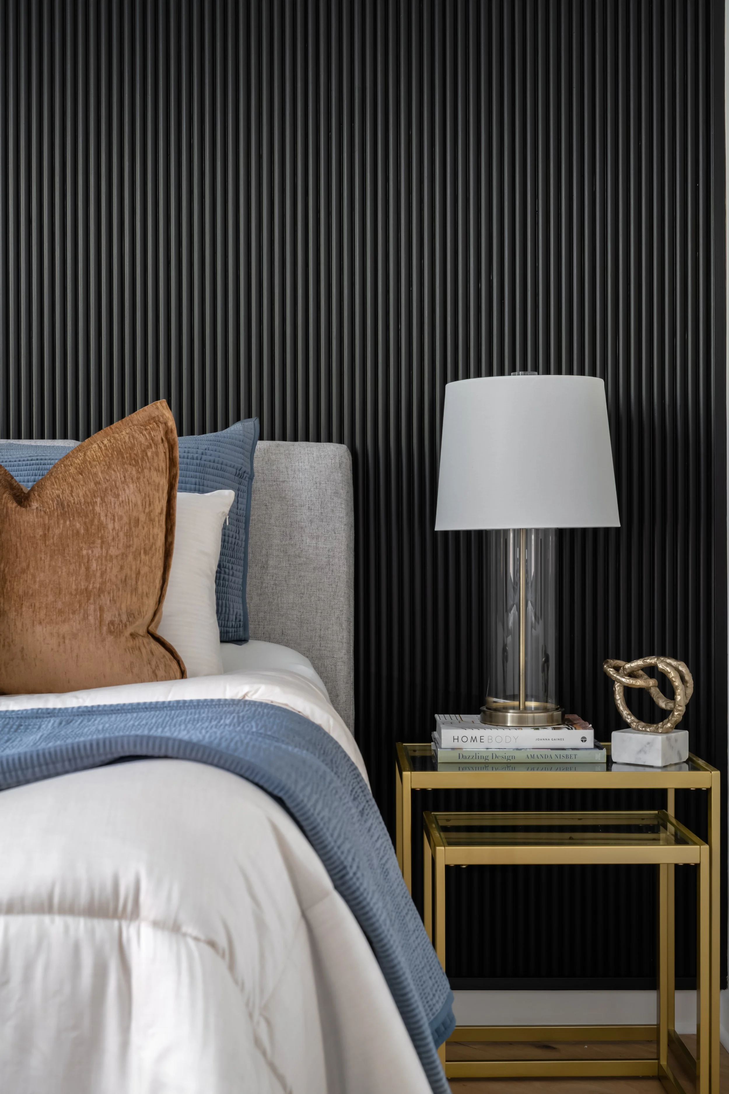 A bedroom corner with a bed, pillows, and a black textured wall. Beside the bed is a gold side table with a stack of books, a white table lamp, and a decorative sculpture.