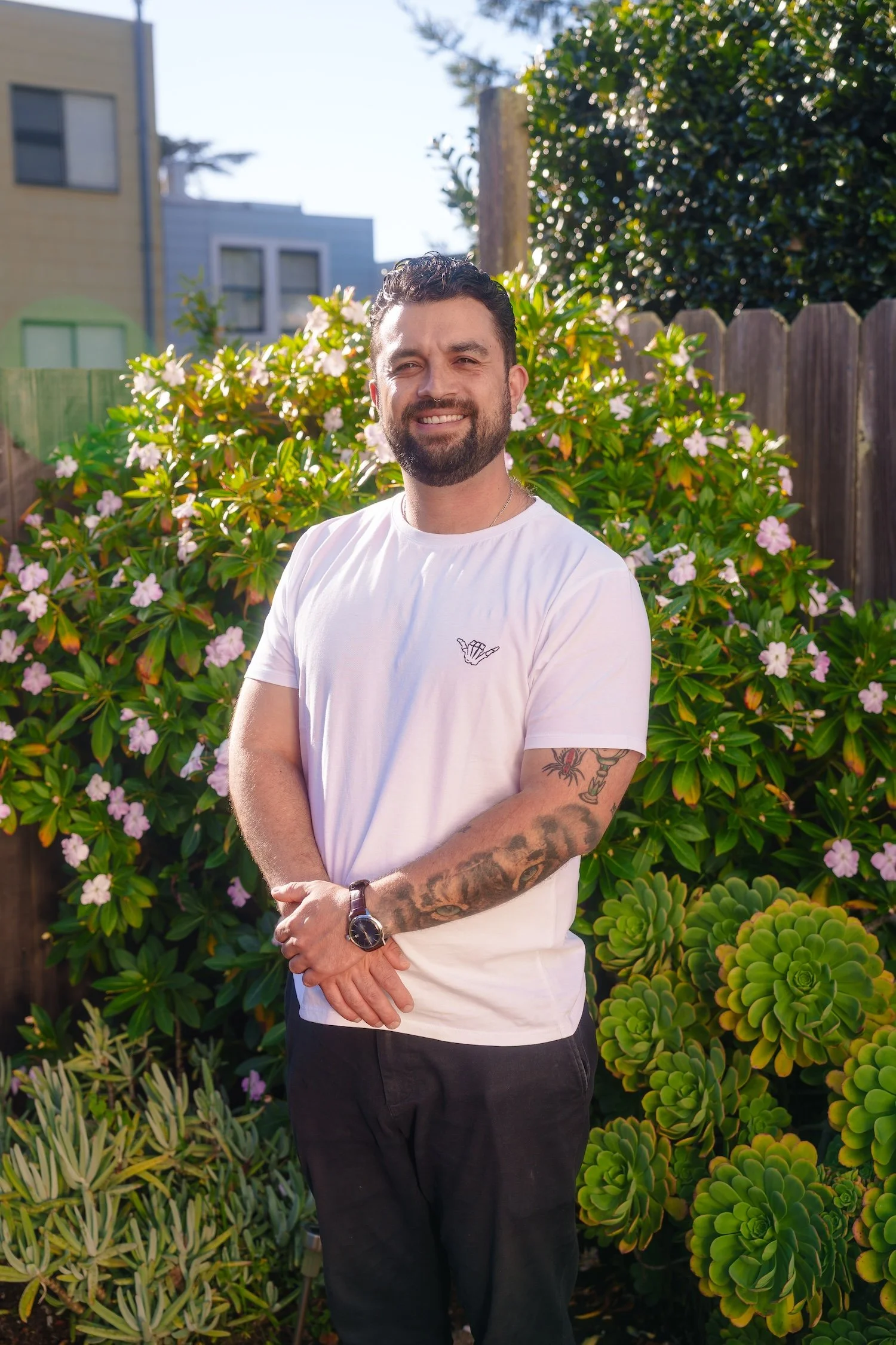 Un hombre de cabello oscuro, barba y tatuajes en el brazo, vestido con camiseta blanca y pantalones oscuros, posando en un jardín con plantas y flores rosadas, bajo la luz del sol.