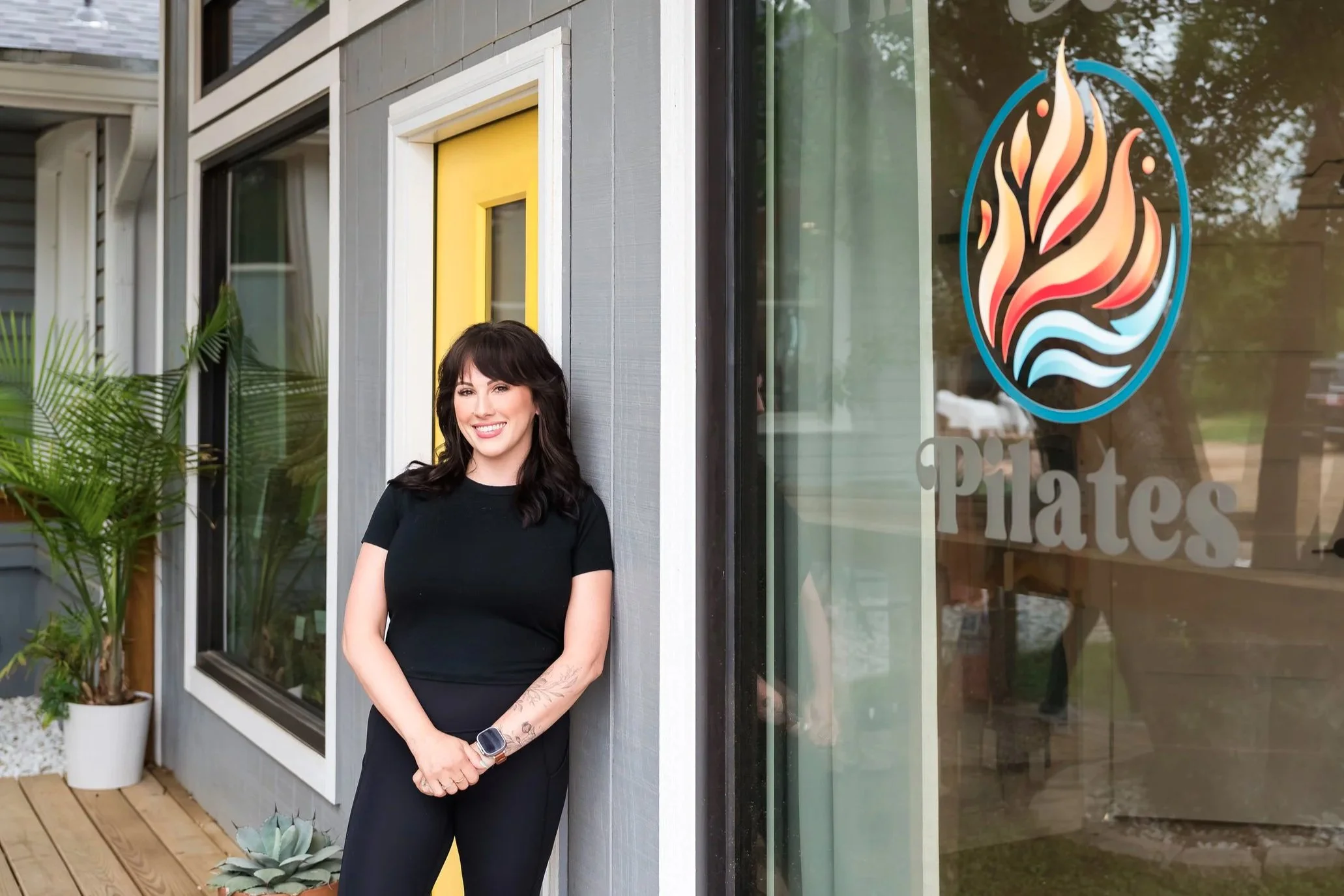 A woman with dark hair and tattoos on her arms, wearing a black t-shirt and a smartwatch, stands outside a building with a large window and a sign that reads 'Pilates.'