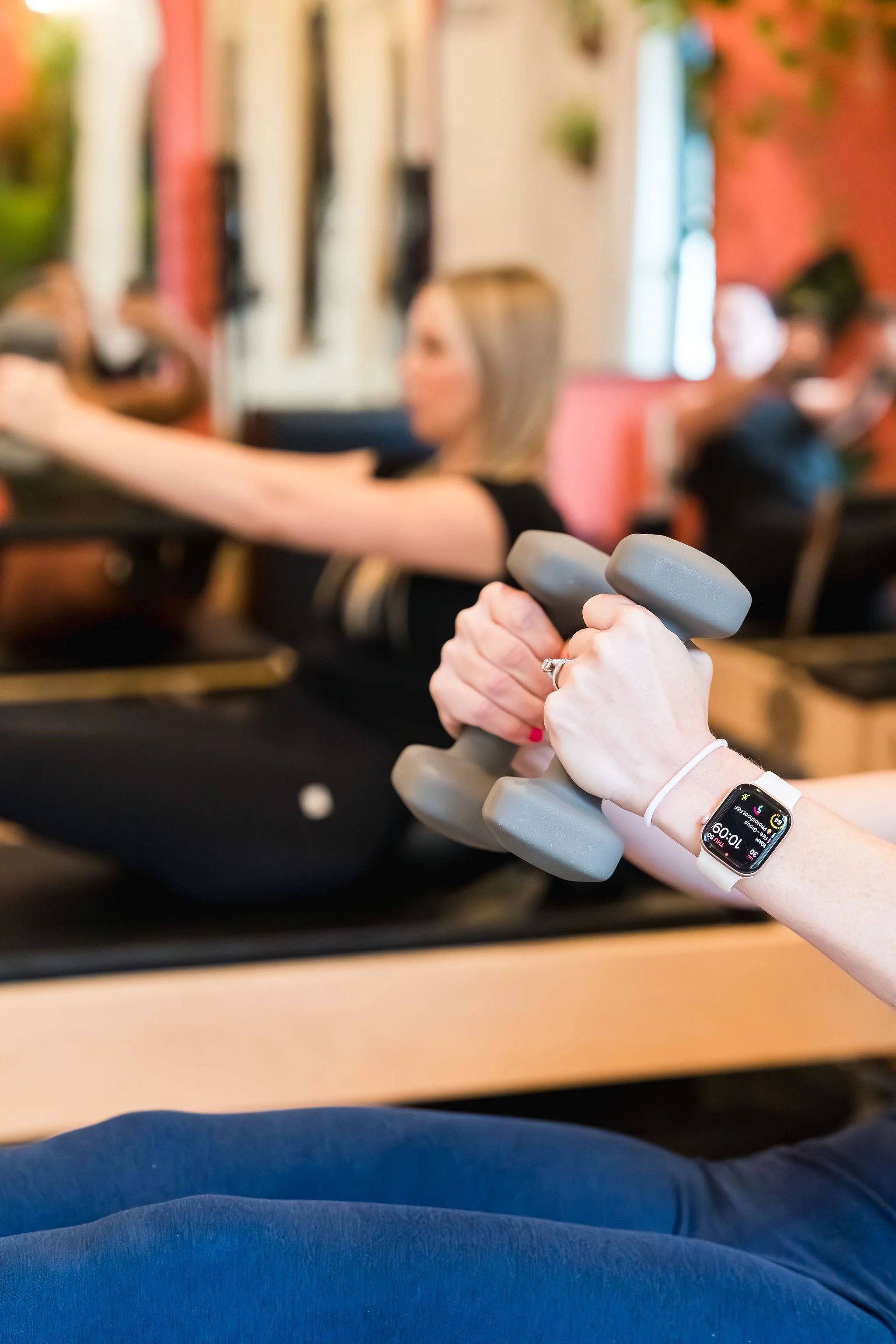 A person holding a gray dumbbell in a fitness class or gym. The focus is on the dumbbell, and a smartwatch with multiple colors and data is visible on their wrist. Other people are blurred in the background.