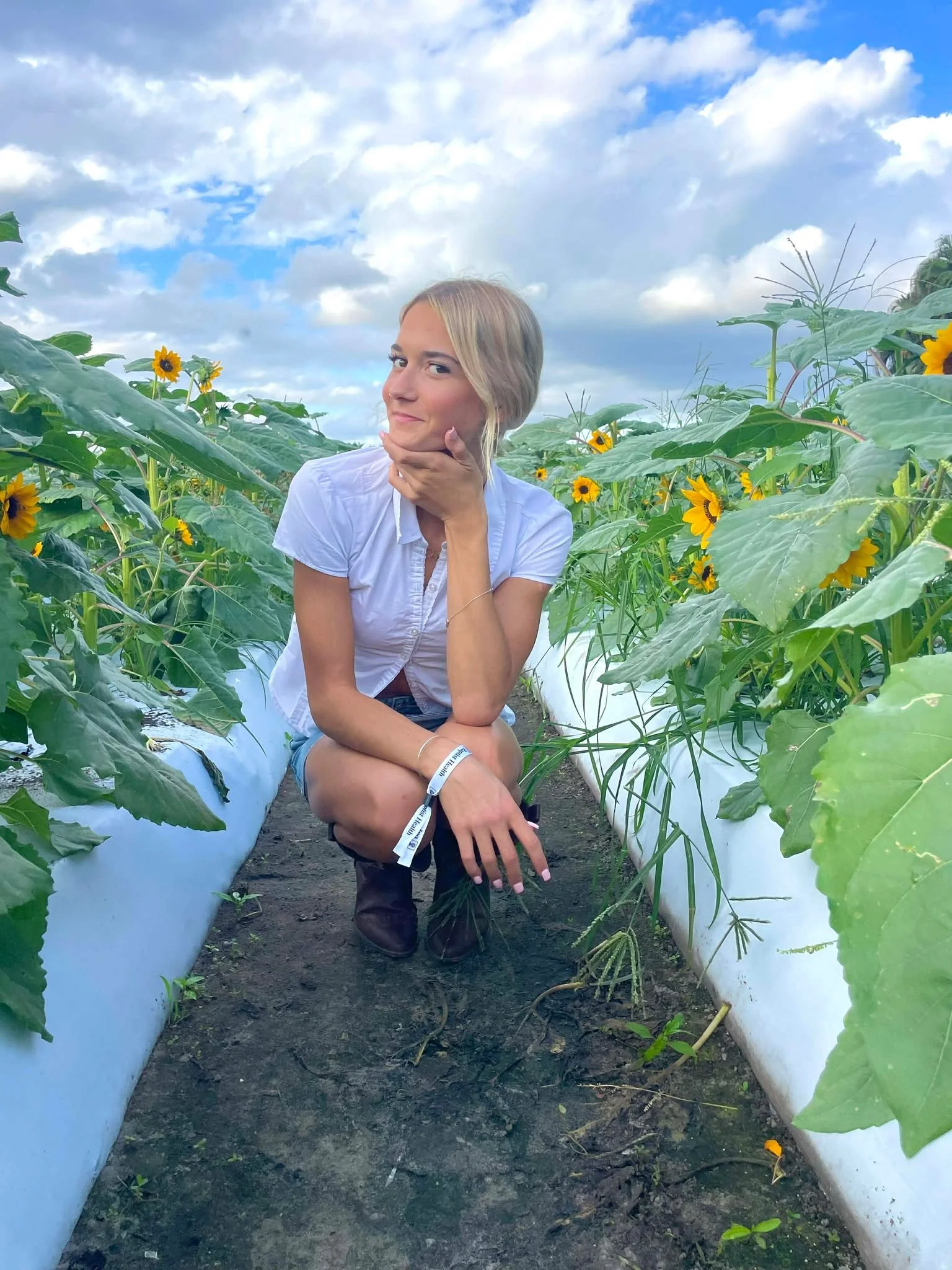 A woman with blonde hair kneeling in a sunflower field, resting her chin on her hand, wearing a white shirt and black boots, with a partly cloudy sky in the background.