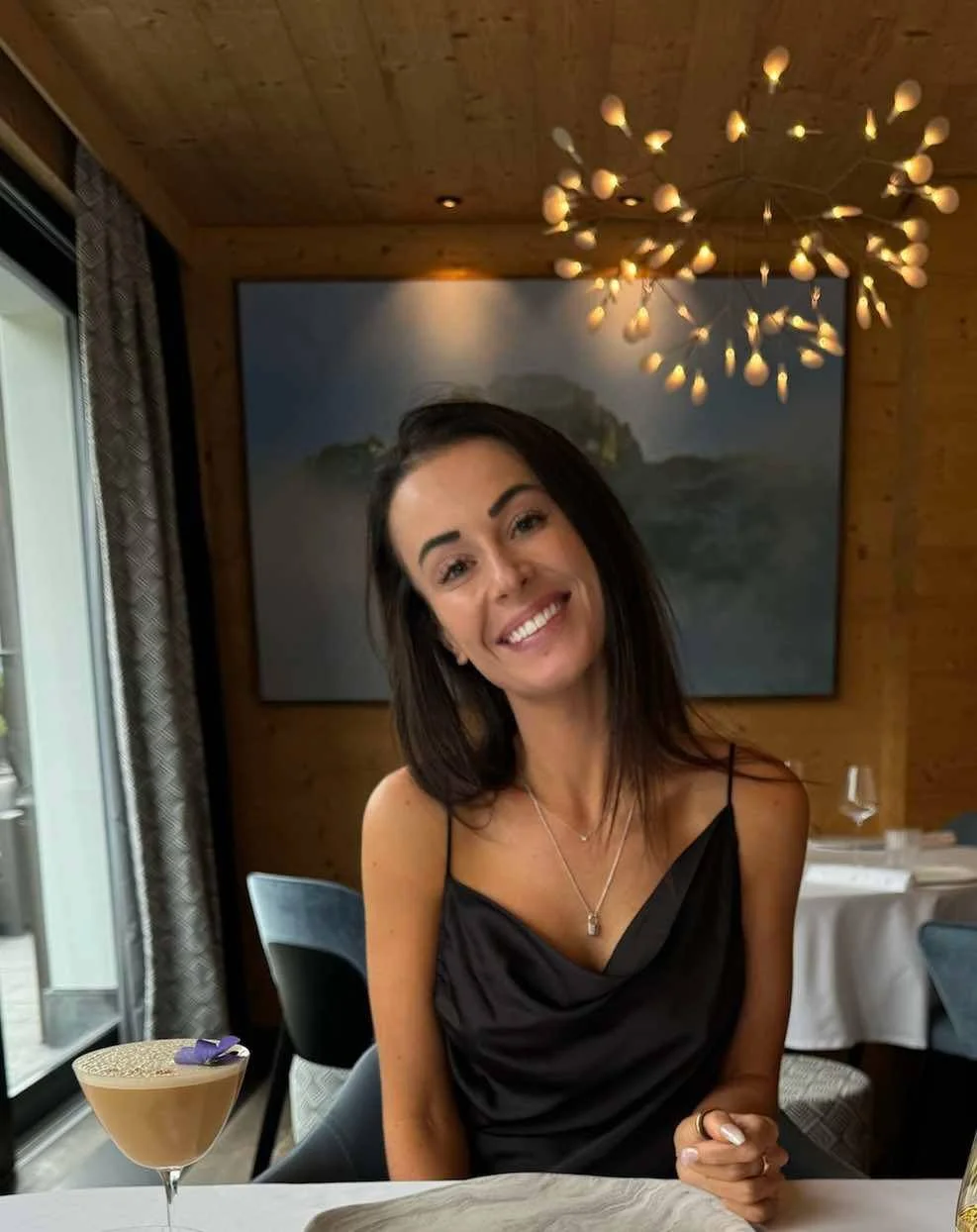 A woman with long dark hair smiling in a restaurant with wooden walls, a large painting, and a modern chandelier. There are tables with white tablecloths and wine glasses.
