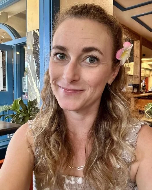 A woman with long curly blonde hair, blue eyes, and a flower in her hair, smiling at the camera inside a cafe or restaurant.