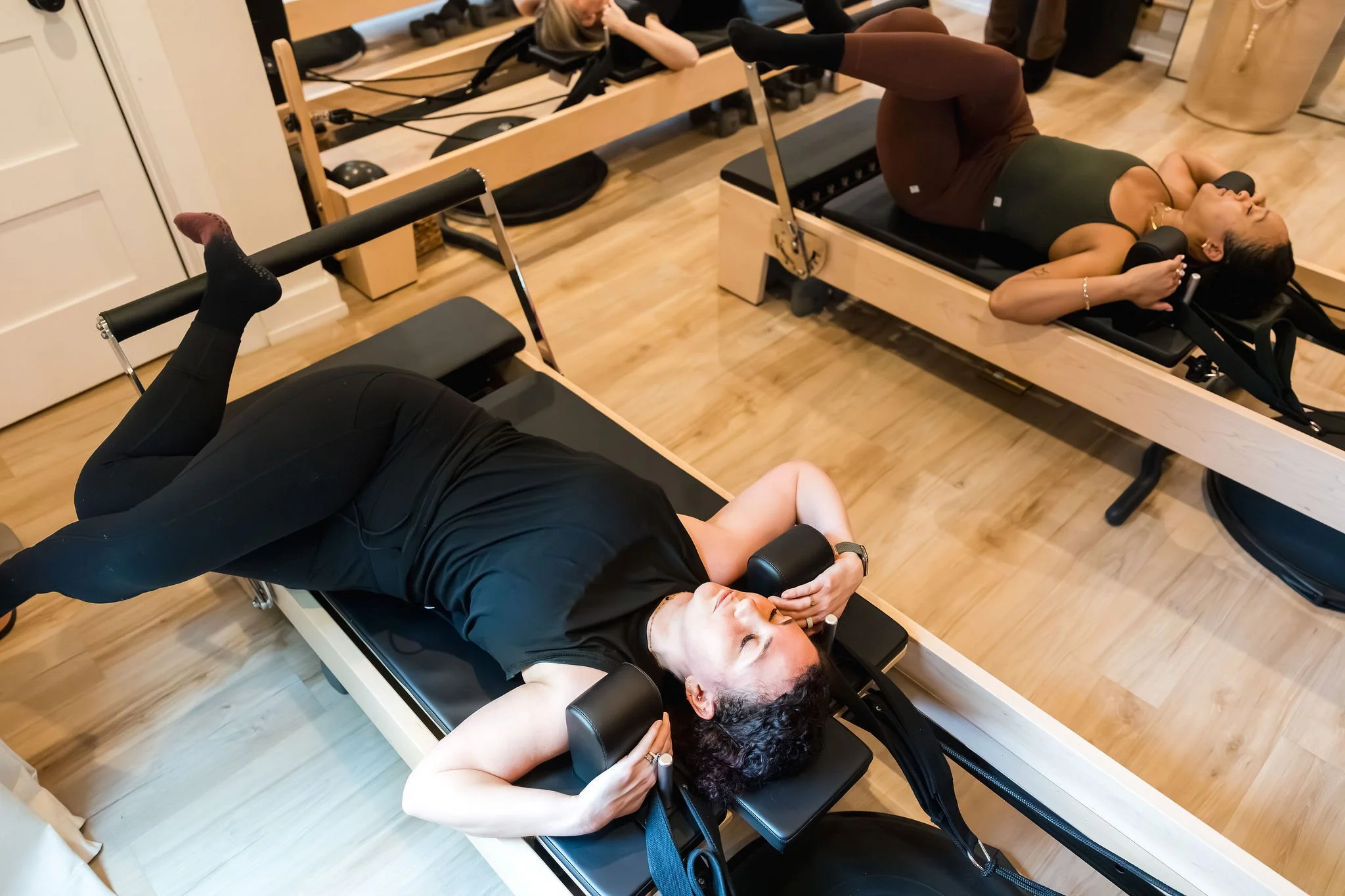 Three women lying on Pilates reformer machines, lifting weights overhead, in a fitness studio with wooden floors and equipment.