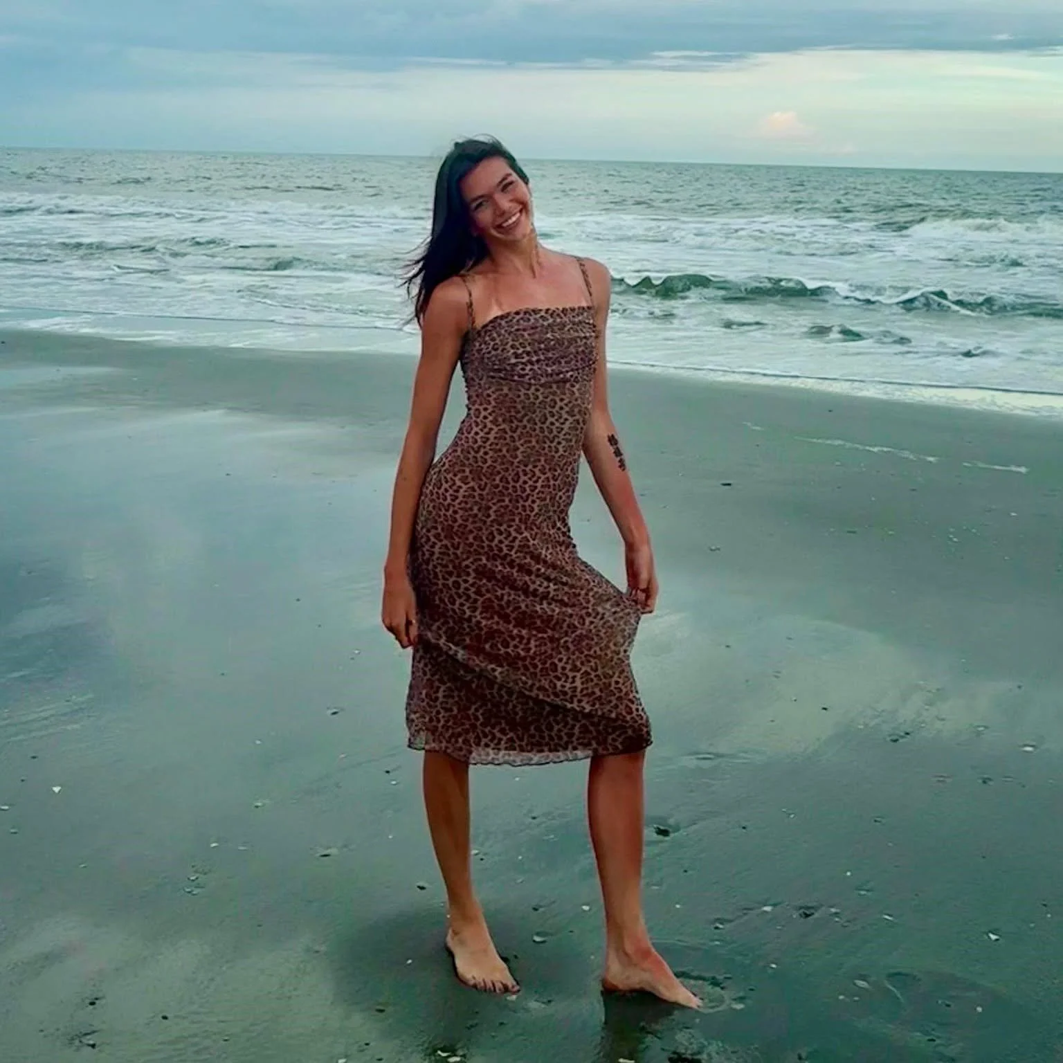 A woman in a leopard print dress smiling and walking barefoot on the beach near the ocean during daytime.