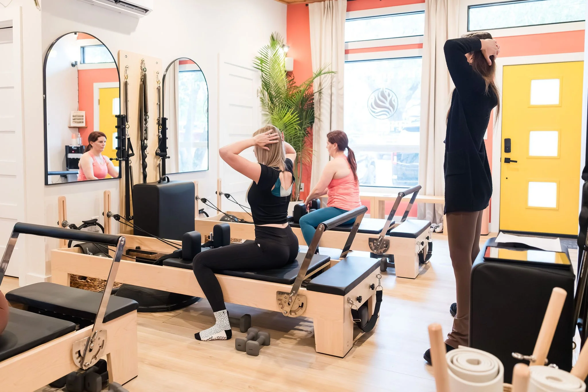 A Pilates studio with several women exercising on Reformers, mirrors on the wall, and a large window with sunlight. One woman is stretching her head back, another is sitting on a reformer, and a staff member or instructor is standing nearby.