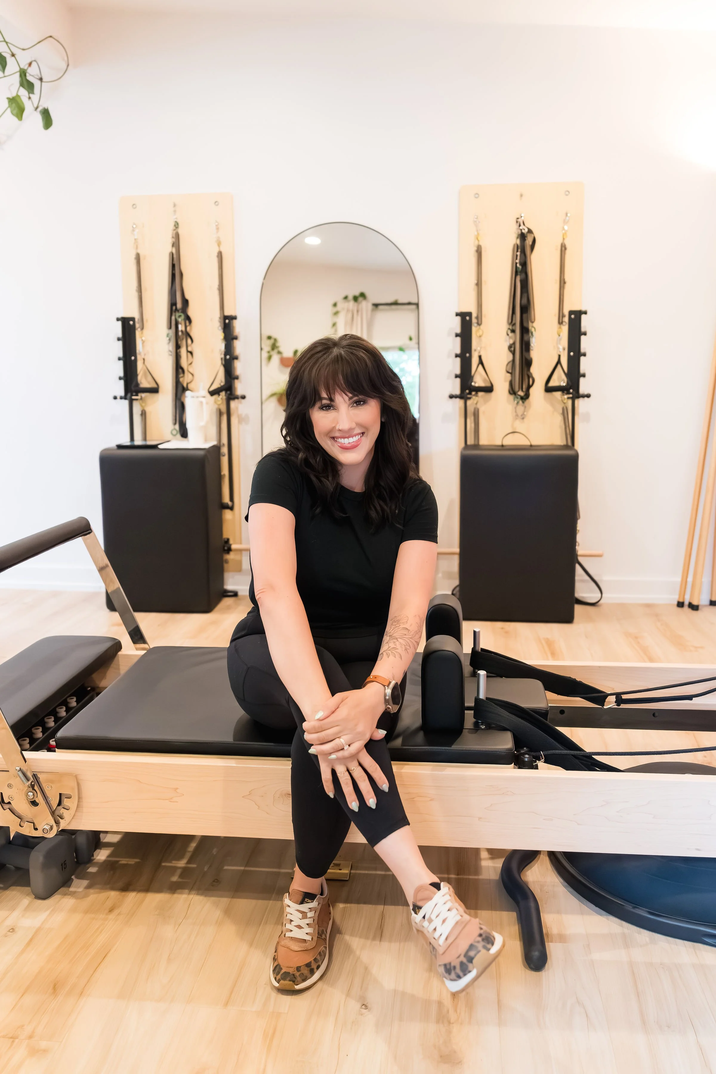 A woman sitting on a Pilates reformer machine in a fitness studio, smiling at the camera. The studio has wooden floors, a mirror, and fitness equipment including resistance bands and mats.