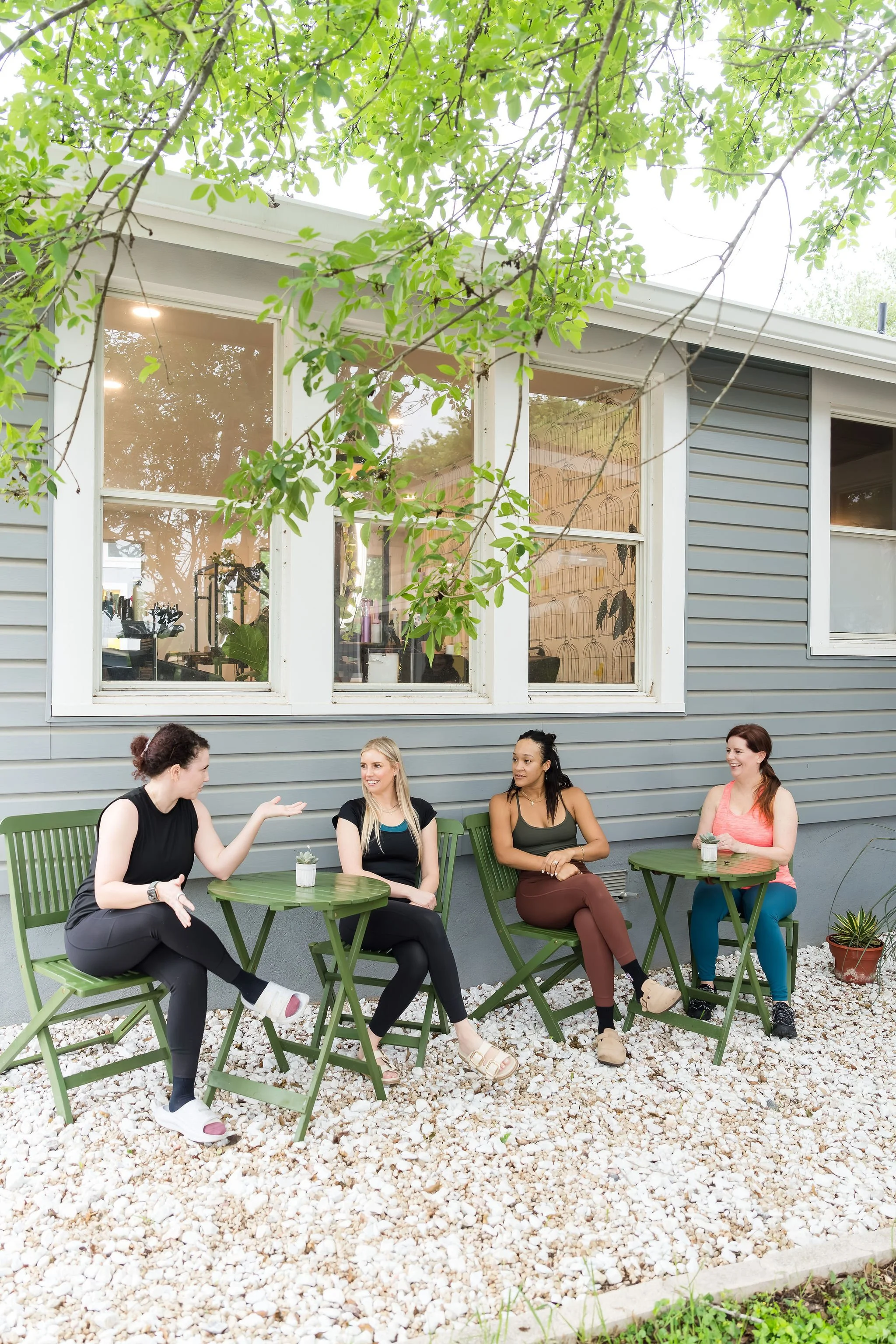Four women sitting on green outdoor chairs at round tables in a backyard, engaging in conversation. The yard has white gravel flooring, and a house with blue siding and large windows is in the background, partially covered by tree branches and leaves.