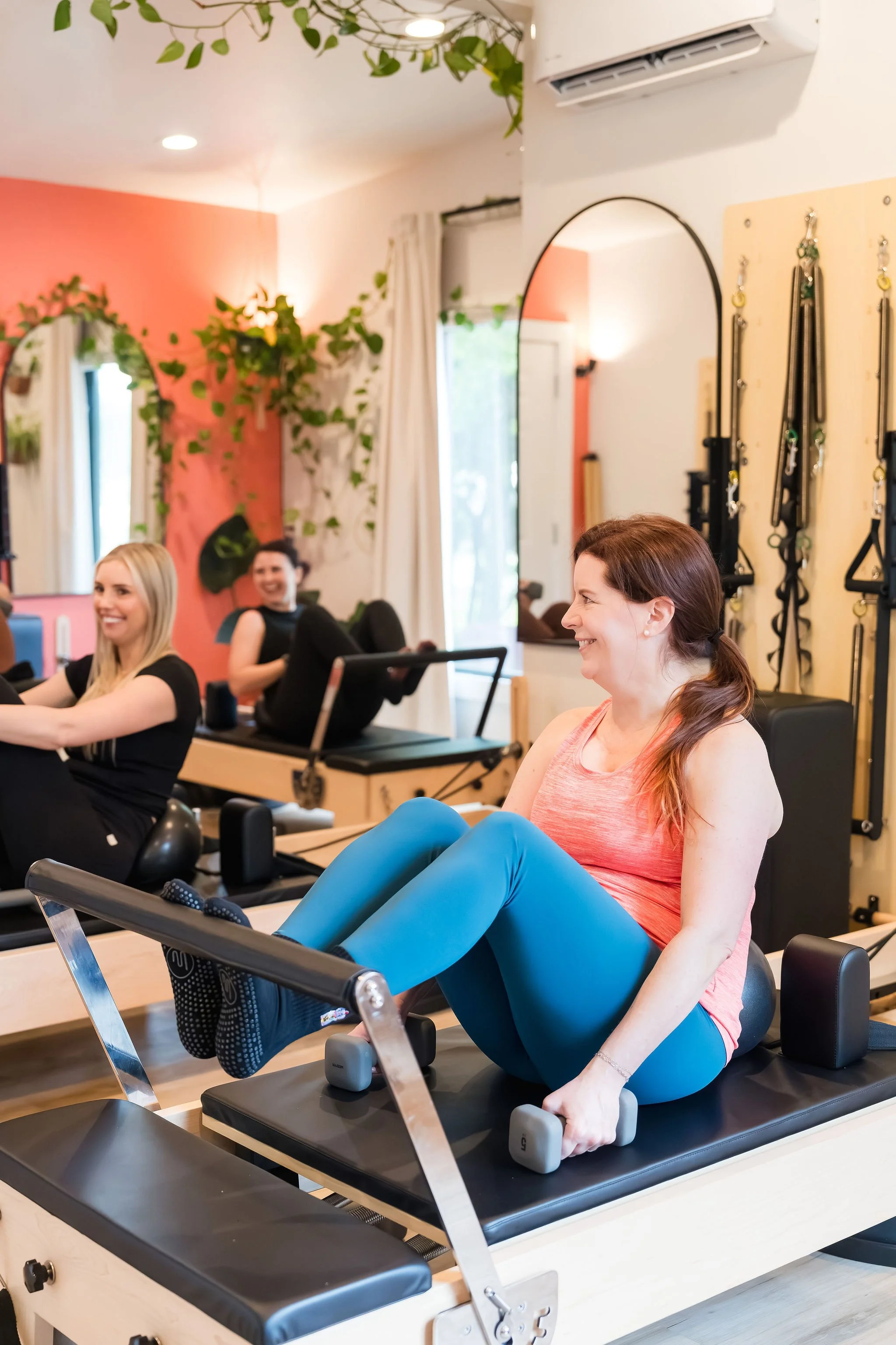 Women participating in a Pilates class using reformer machines in a bright, airy gym with plants and mirrors.