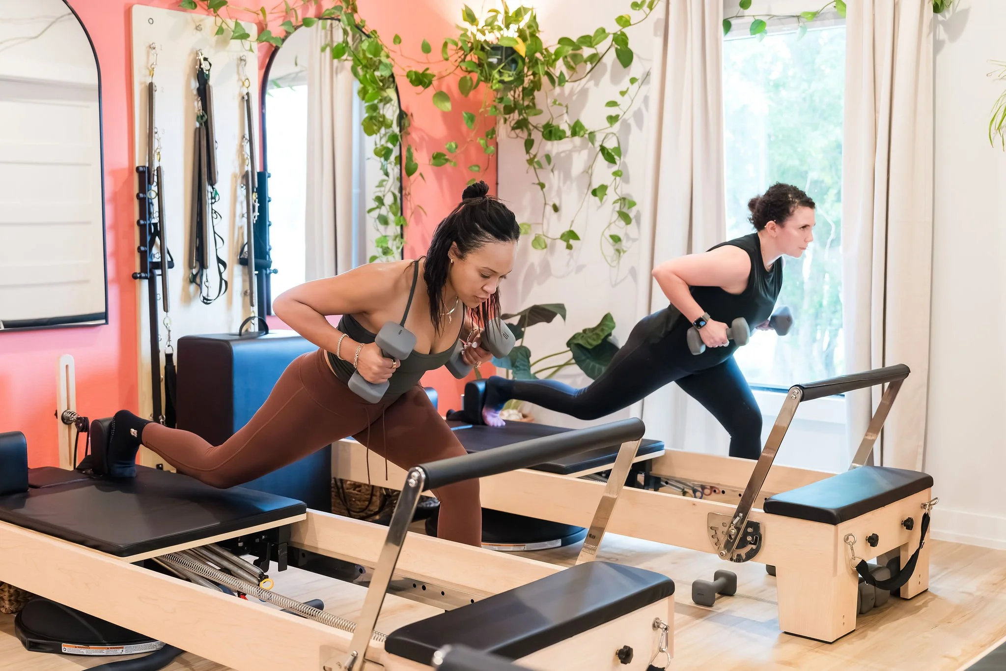 Two women are exercising on Pilates reformer machines in a bright room with green plants, a pink accent wall, and large windows with white curtains.