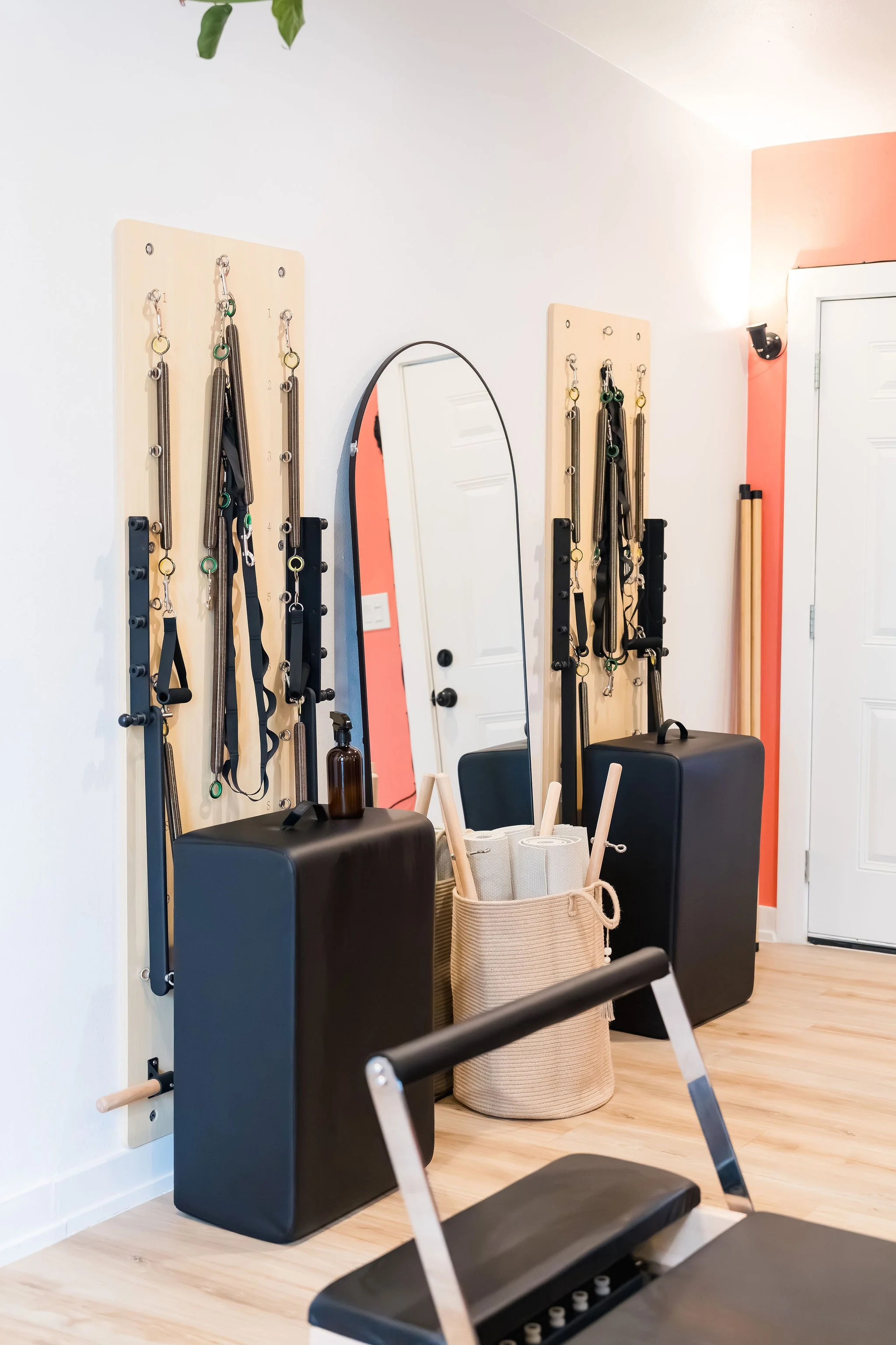 Interior view of a room with wall-mounted racks holding resistance bands, a mirror, a black stool, and a woven basket containing rolled towels and wooden massage rollers. The room features light-colored wooden flooring and a corner wall painted in coral pink.