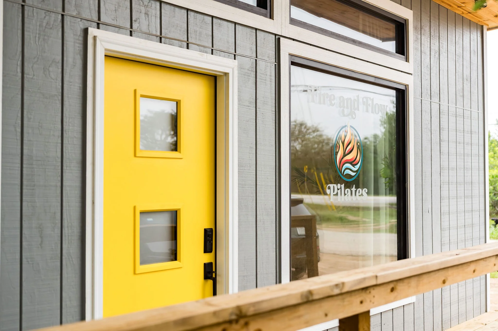 Close-up of a modern building's exterior with gray wooden panel siding, large windows, and a bright yellow door with small square windows. There is a sign on the window that reads 'Fire and Flow' with a colorful flame logo above the text 'Pilates.'