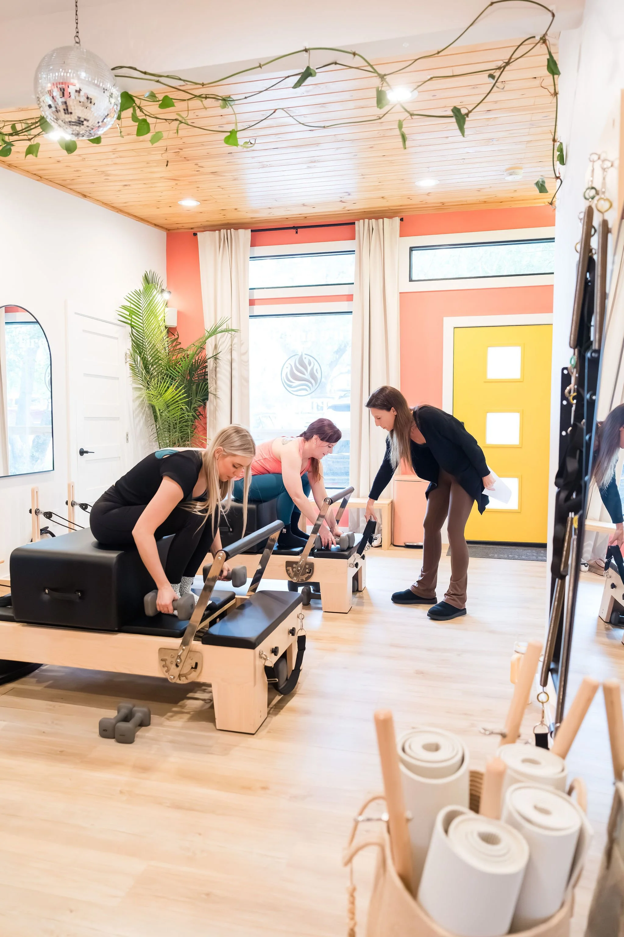 Four women are exercising on Pilates reformer machines in a brightly lit room with a wooden ceiling. There are large windows with curtains, a yellow door, a tall potted plant, and a mirror on the wall. In the foreground, there is a basket filled with rolled-up white yoga mats and some wooden sticks.
