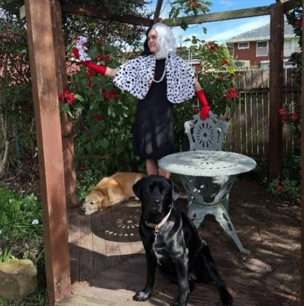 A woman dressed as Cruella de Vil, wearing a black dress with a white and black spotted cape, red gloves, and a white wig, standing under a wooden pergola with two dogs in a garden. One dog is black, sitting on the deck, and the other is golden, lying down.