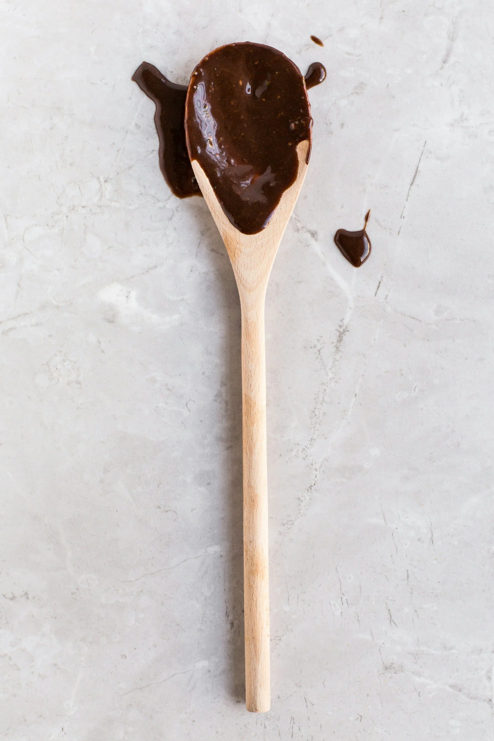 A wooden spoon with melted chocolate on top, with some melted chocolate spilled on a light-colored surface.
