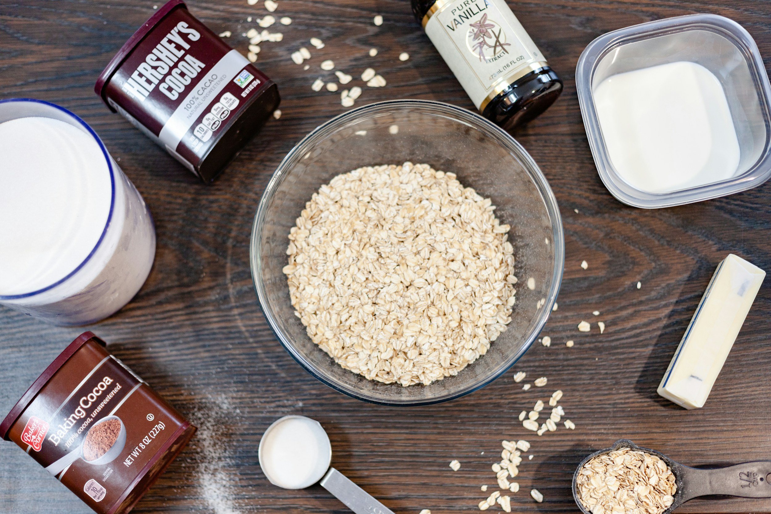 Oats, chocolate, vanilla, and dairy ingredients on a wooden surface, possibly ingredients for a recipe.
