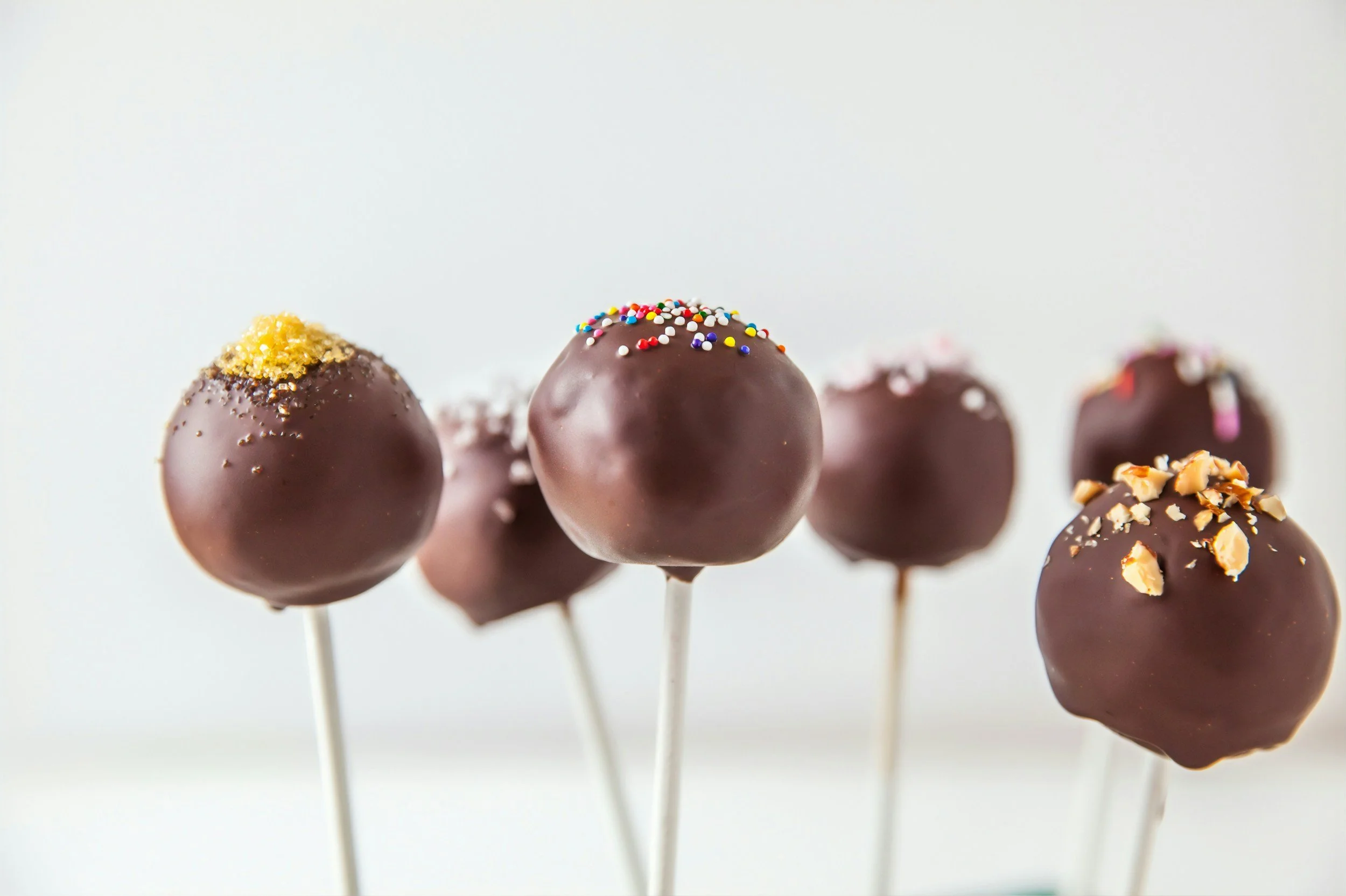 Six chocolate cake pops decorated with sprinkles and crushed nuts on white sticks against a white background.