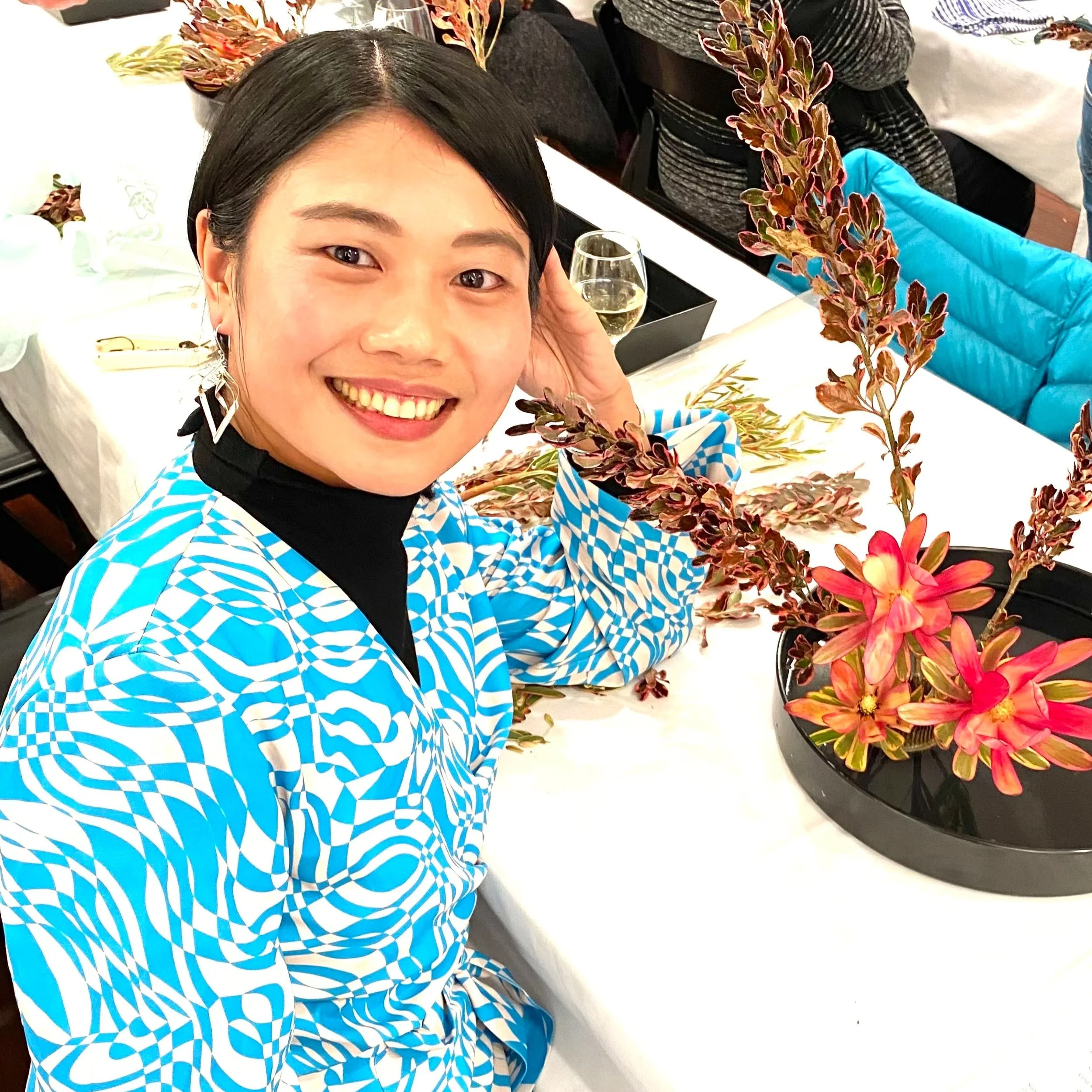 A smiling woman with black hair and earrings, wearing a blue and white checkered shirt, sitting at a table with red and green potted plants and a glass of champagne.