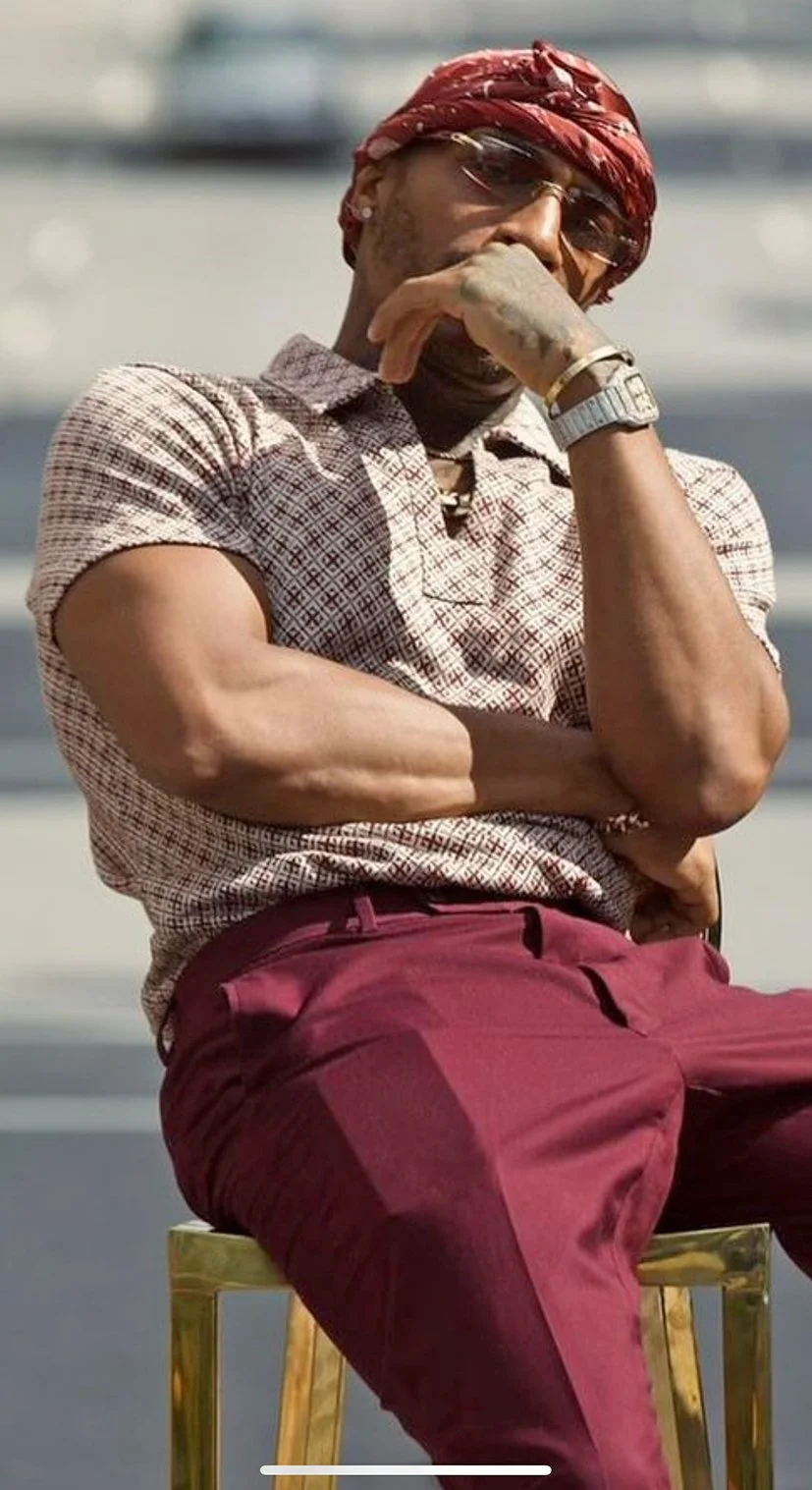 A man sitting on a gold chair outdoors wearing a burgundy scarf, glasses, and a checkered short-sleeve shirt, resting his chin on his hand with his arm crossed.