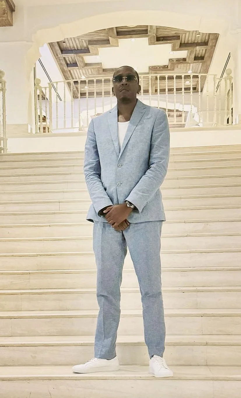 A man in a light gray suit with white sneakers, wearing sunglasses, stands on a grand staircase in a well-lit, elegant interior.