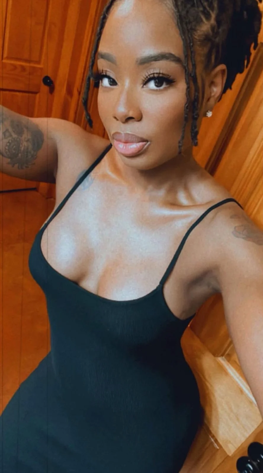 A woman with tattoos on her arms taking a selfie in a room with wooden walls, wearing a black spaghetti strap dress, with her hair styled in braids.
