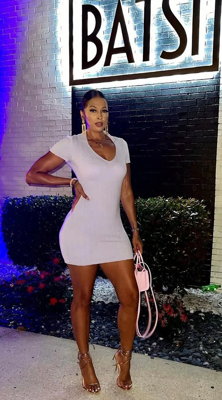 A woman in a short white dress standing in front of a black brick wall with a sign that reads 'BATSI'. She is holding a pink purse and wearing high-heeled sandals, jewelry, and makeup.