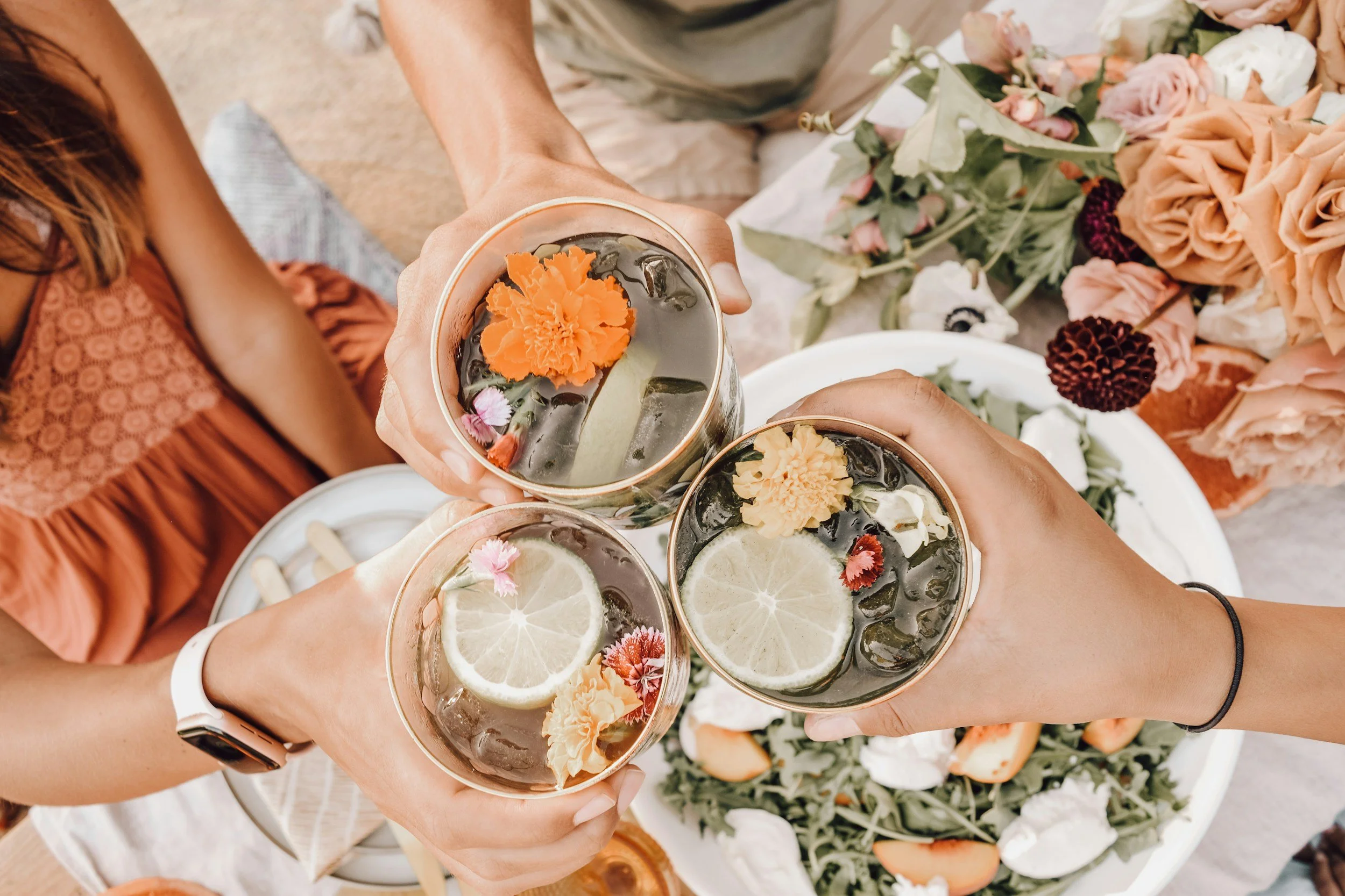 Three hands holding cocktails with flowers in them cheersing above view