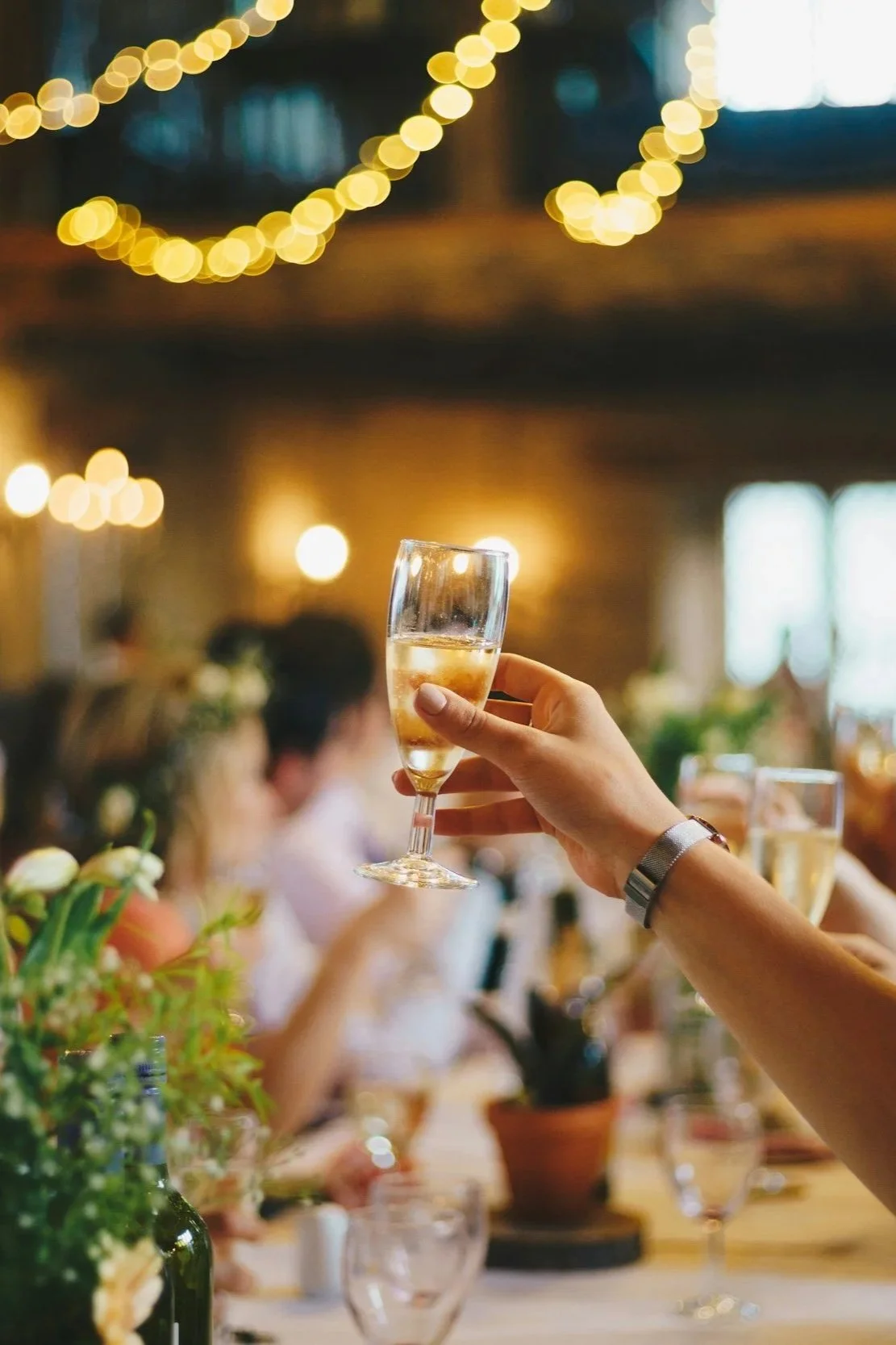 A person holding a glass of champagne at a social gathering, with other people sitting at a table in the background and string lights overhead.