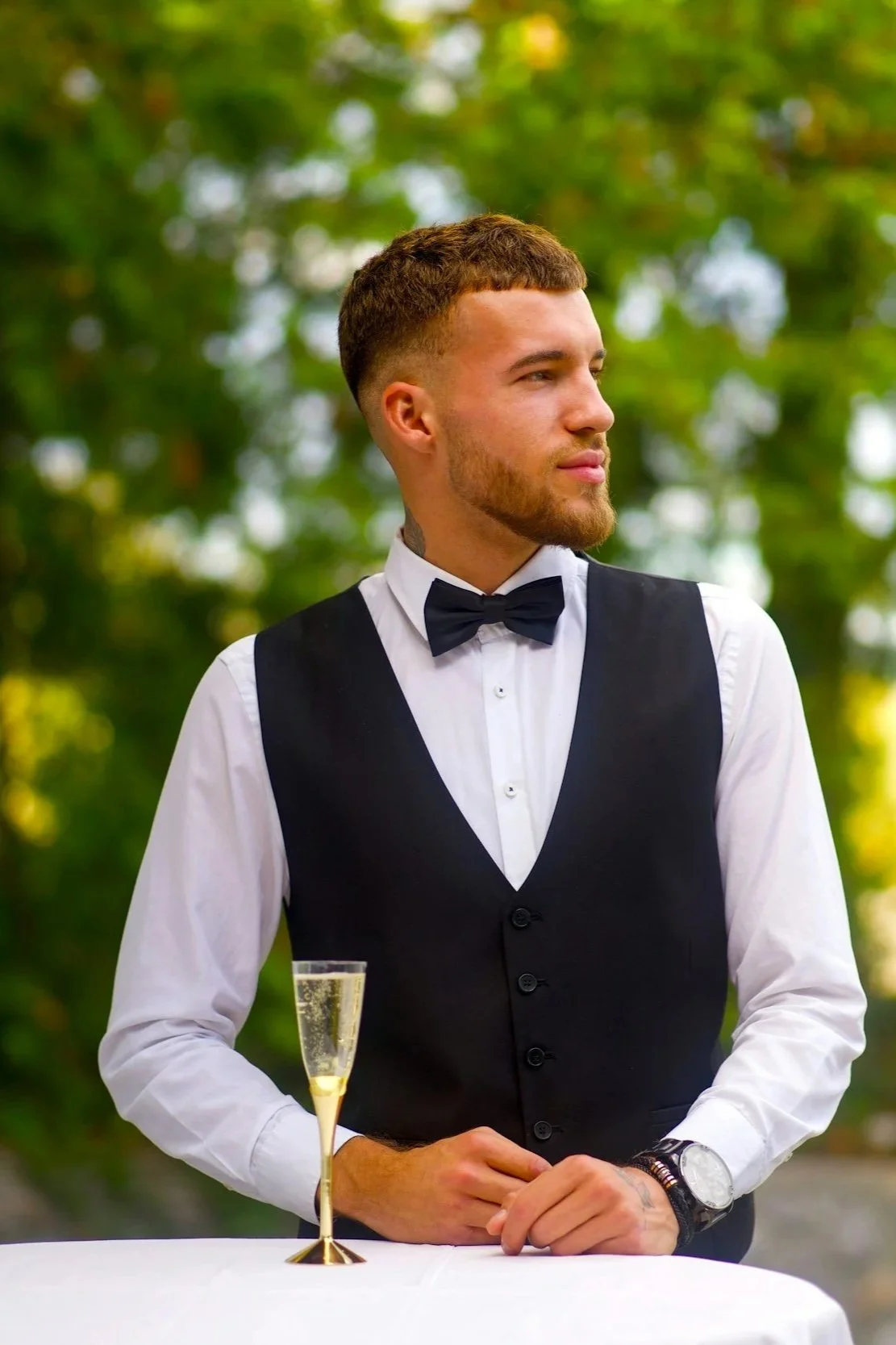 Server in vest and tie standing outside near table with champagne flute on it blurred background
