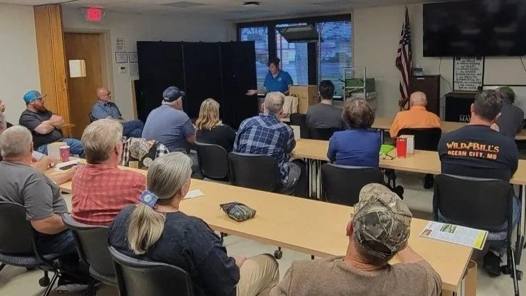 A group of people attending a meeting or presentation in a room, seated at tables, listening to a speaker standing at the front near a large window, with an American flag and a TV on the wall nearby.