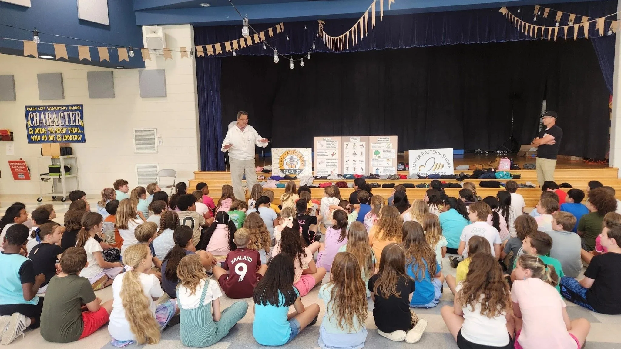 A school assembly with a large group of children sitting on the floor facing a stage where two speakers are talking. The stage has posters and banners about beekeeping and pollination, and the auditorium walls have decorations including banners and signs.