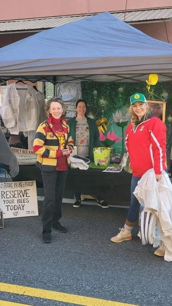 Three women standing behind a booth at an outdoor market, with a blue canopy overhead. The booth has signs about honey bees and reserve bees today. One woman is wearing a red and yellow jacket, another in a red jacket with a green cap, and the third in a gray sweatshirt. There are various items and decor at the booth, including a framed picture and a yellow lantern.