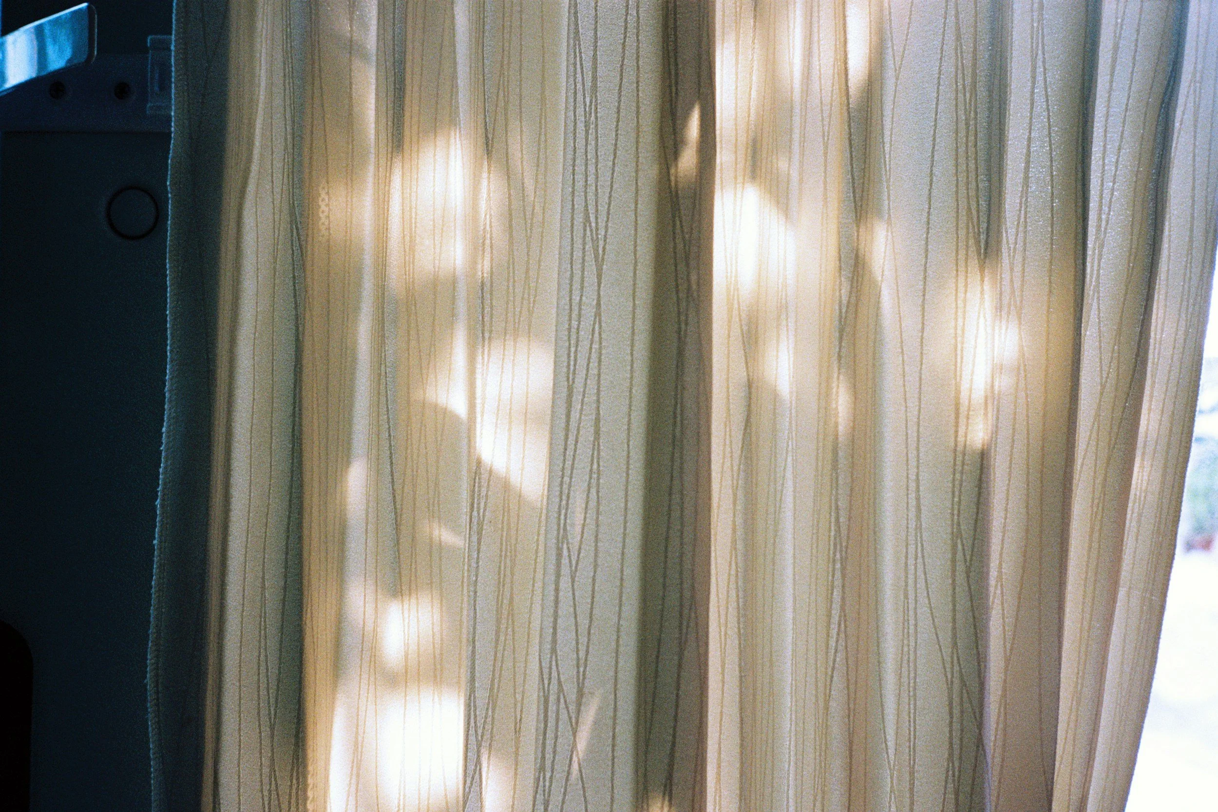 Close-up of a window with beige curtains, sunlight filtering through, creating shadows and light patterns.