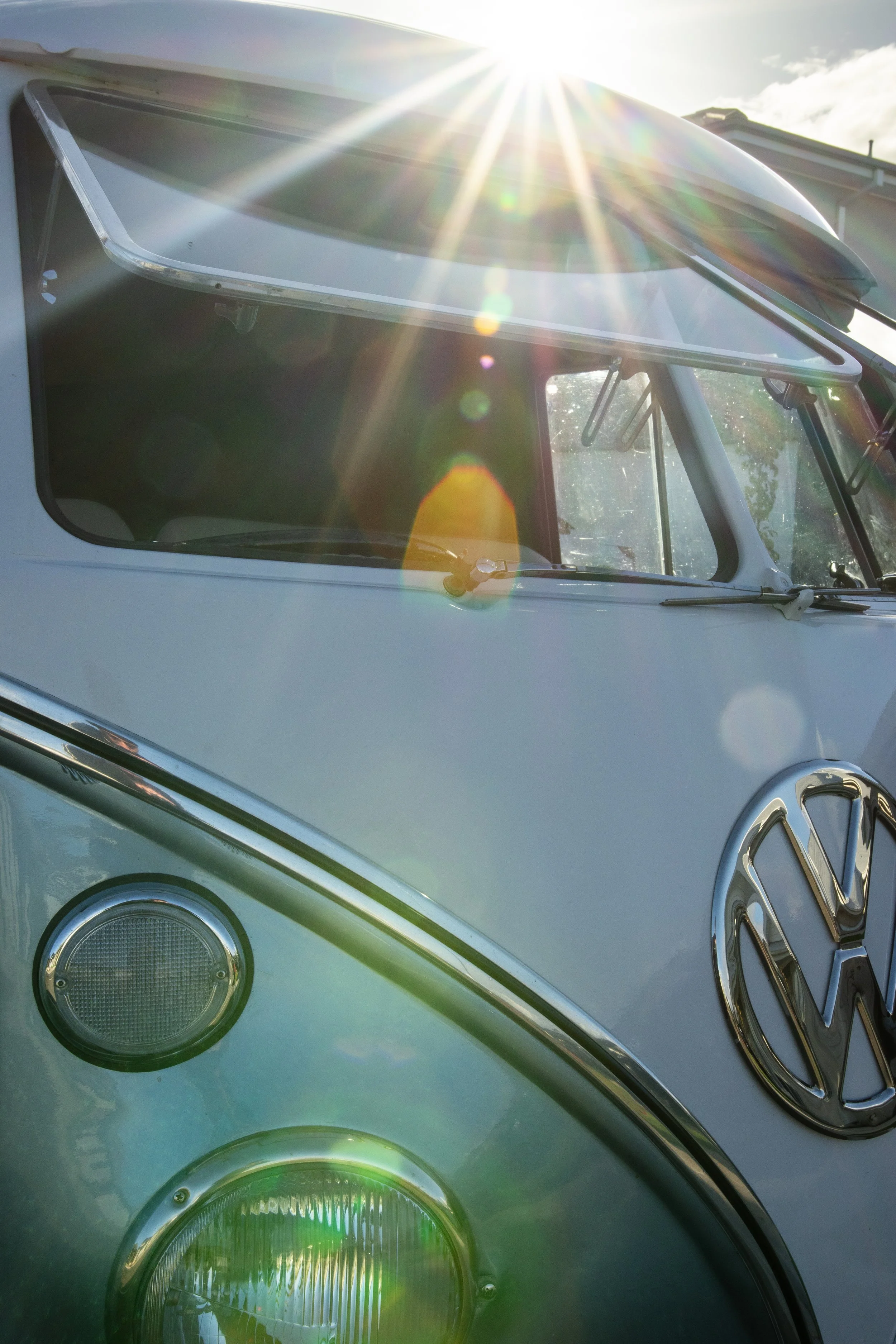 Close-up of a classic Volkswagen van's front with a partially opened sunroof, sunlight creating lens flare effects, and the Volkswagen logo visible on the front.