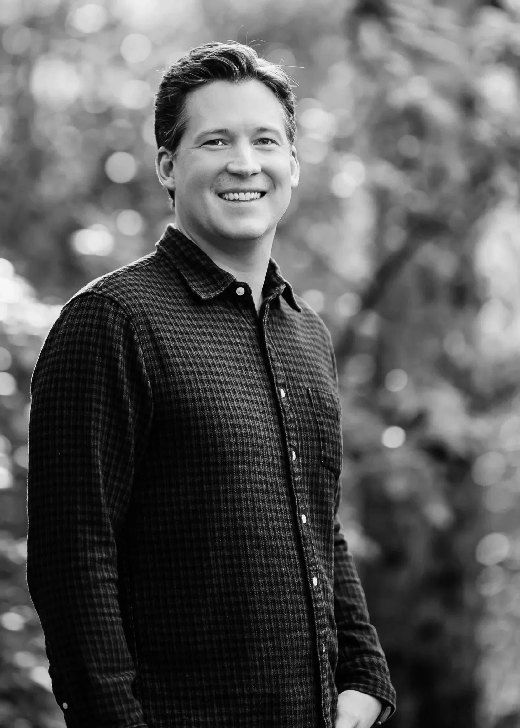 A smiling young man with short dark hair, wearing a checkered shirt, standing outdoors with a blurred background of trees.