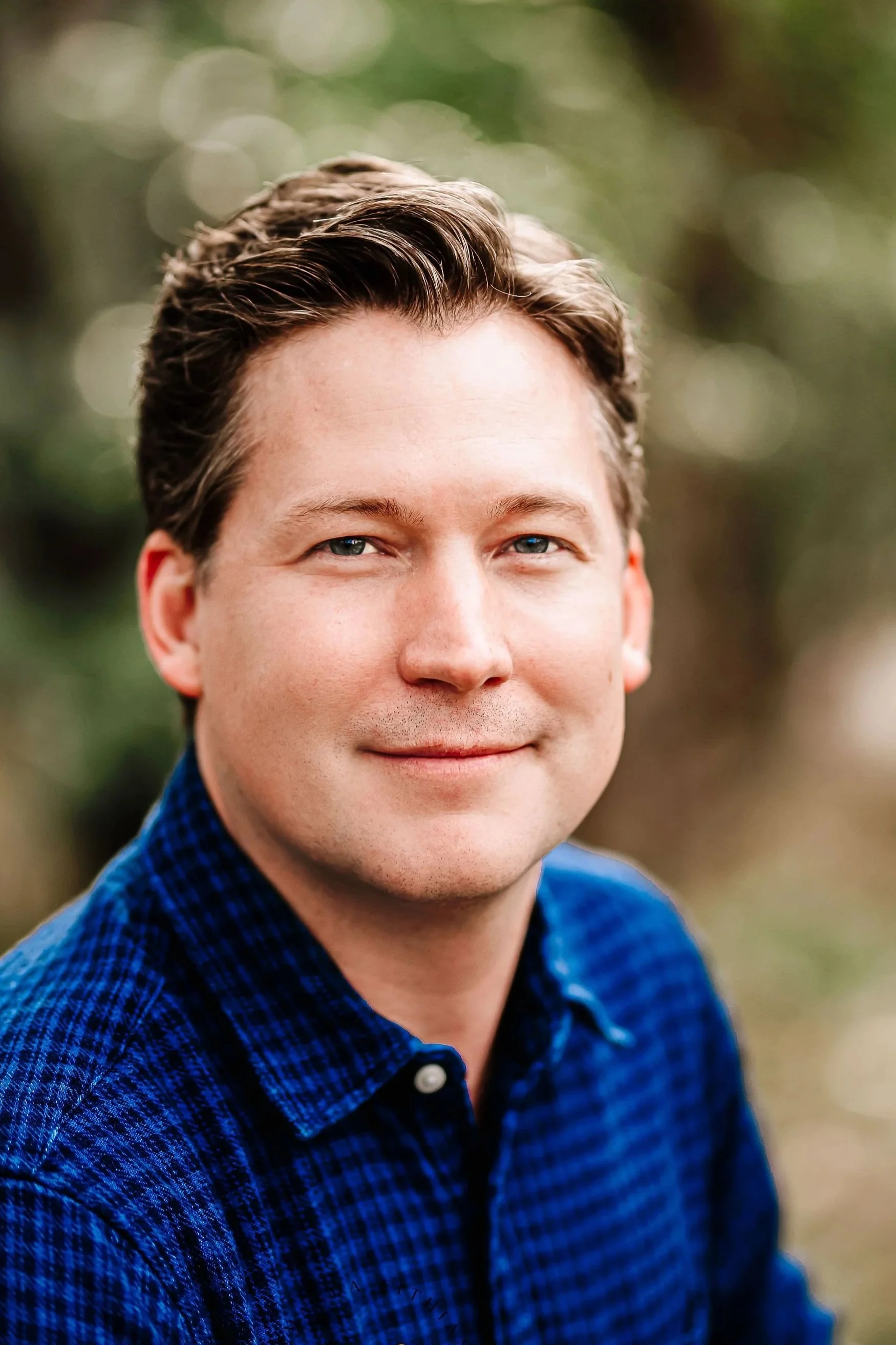 Close-up of a smiling man with short, wavy brown hair, blue eyes, wearing a dark blue plaid shirt, outdoor background with blurred trees and greenery.