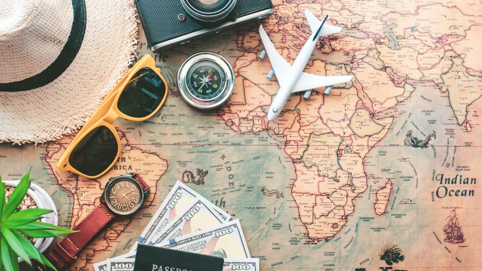 World travel planning items placed on a map of Africa, including a hat, sunglasses, a wristwatch, a toy airplane, a camera, a compass, a model airplane, dollar bills, and a passport.