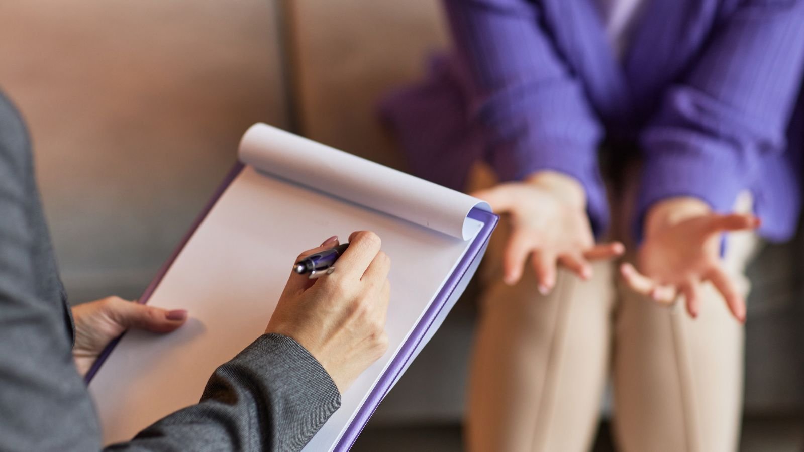 A person in a gray jacket is writing on a notepad with a black pen. Another person in a purple shirt and beige pants is gesturing with open hands, with a blurred background.