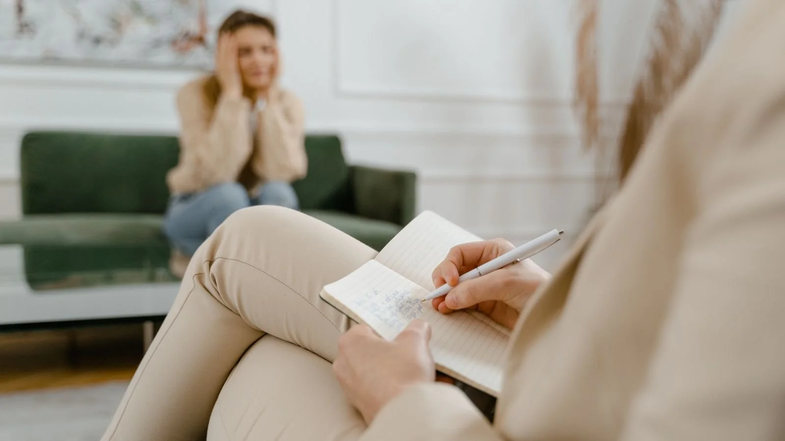 A person in beige clothing taking notes in a notebook with a pen, while a woman with hands on her face sits on a green couch in the background, appearing distressed.