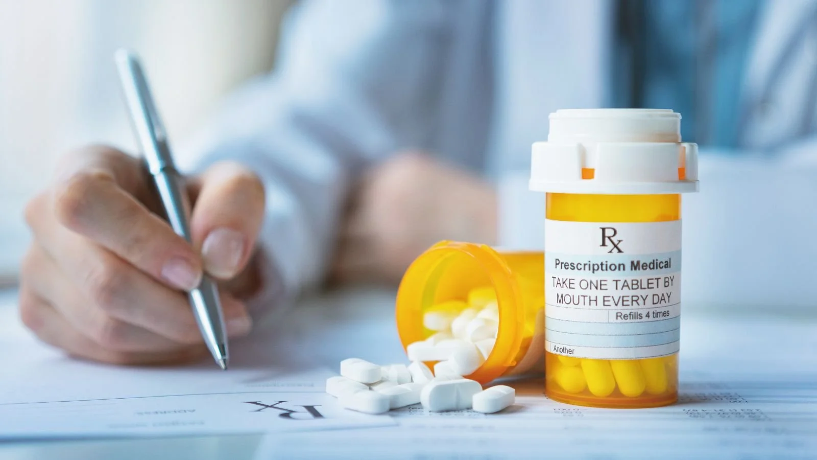 A person writing on a prescription paper with a pen, with a prescription medication bottle and spilled pills on the table.